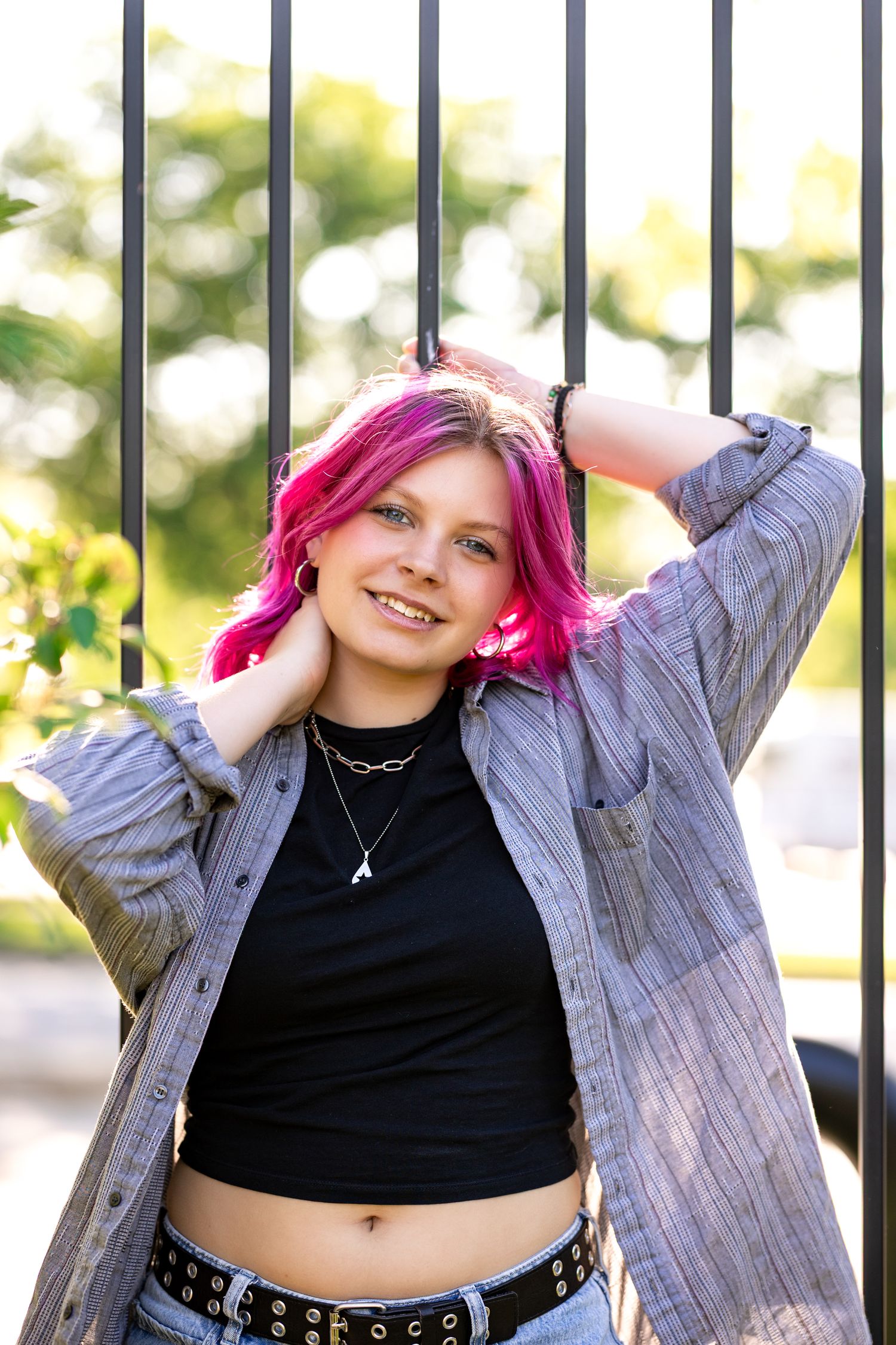 Young woman with pink hair smiling, wearing a black crop top and gray shirt, posing outdoors near a metal fence.