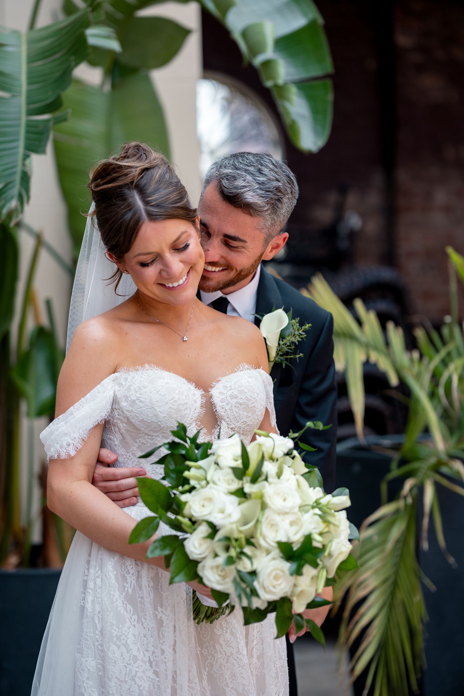 Bride and groom embracing outdoors, bride holding white rose bouquet in lace off-shoulder gown with veil, groom in dark suit.