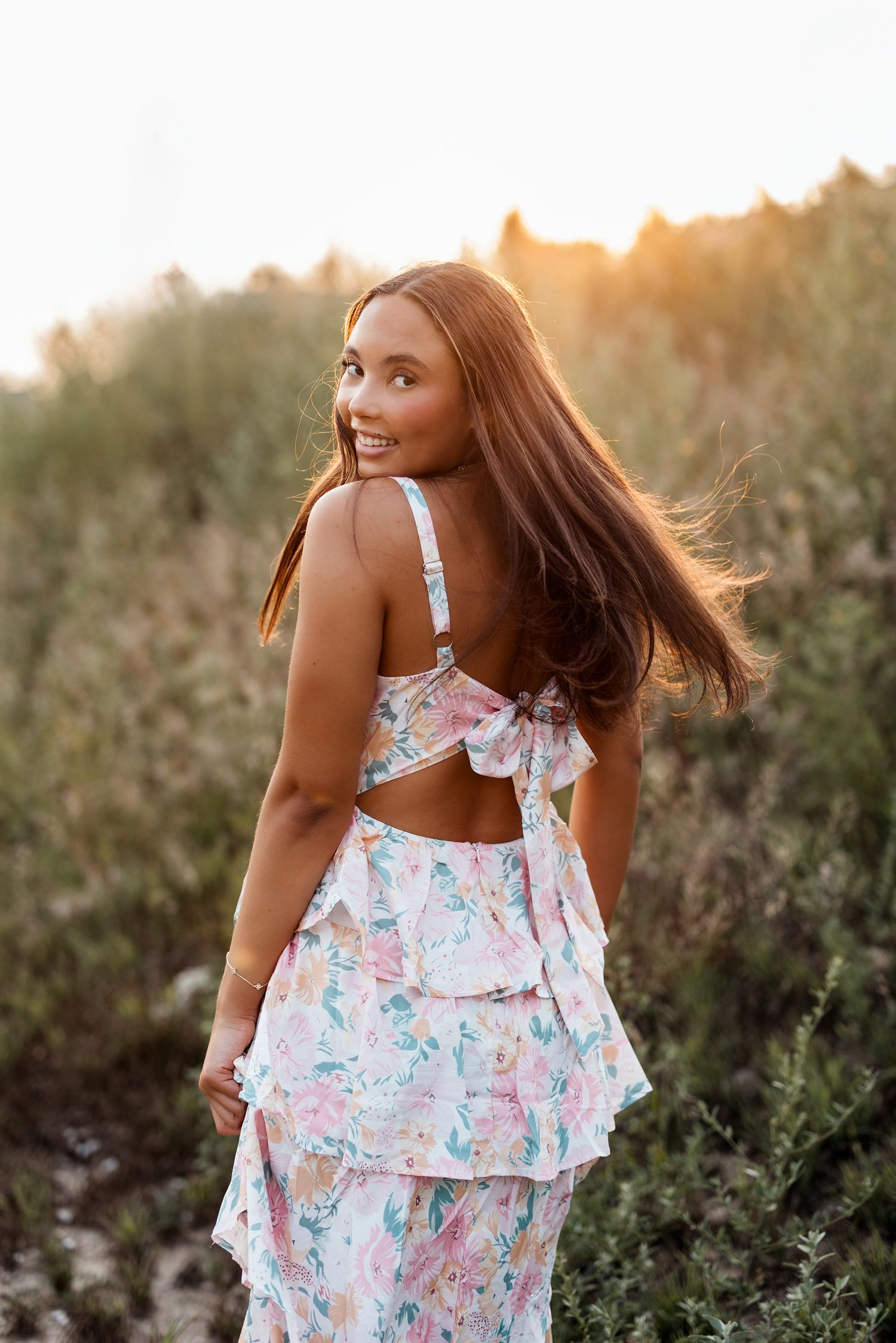 Young woman in floral ruffle dress smiling over shoulder at golden sunset in a natural field setting.