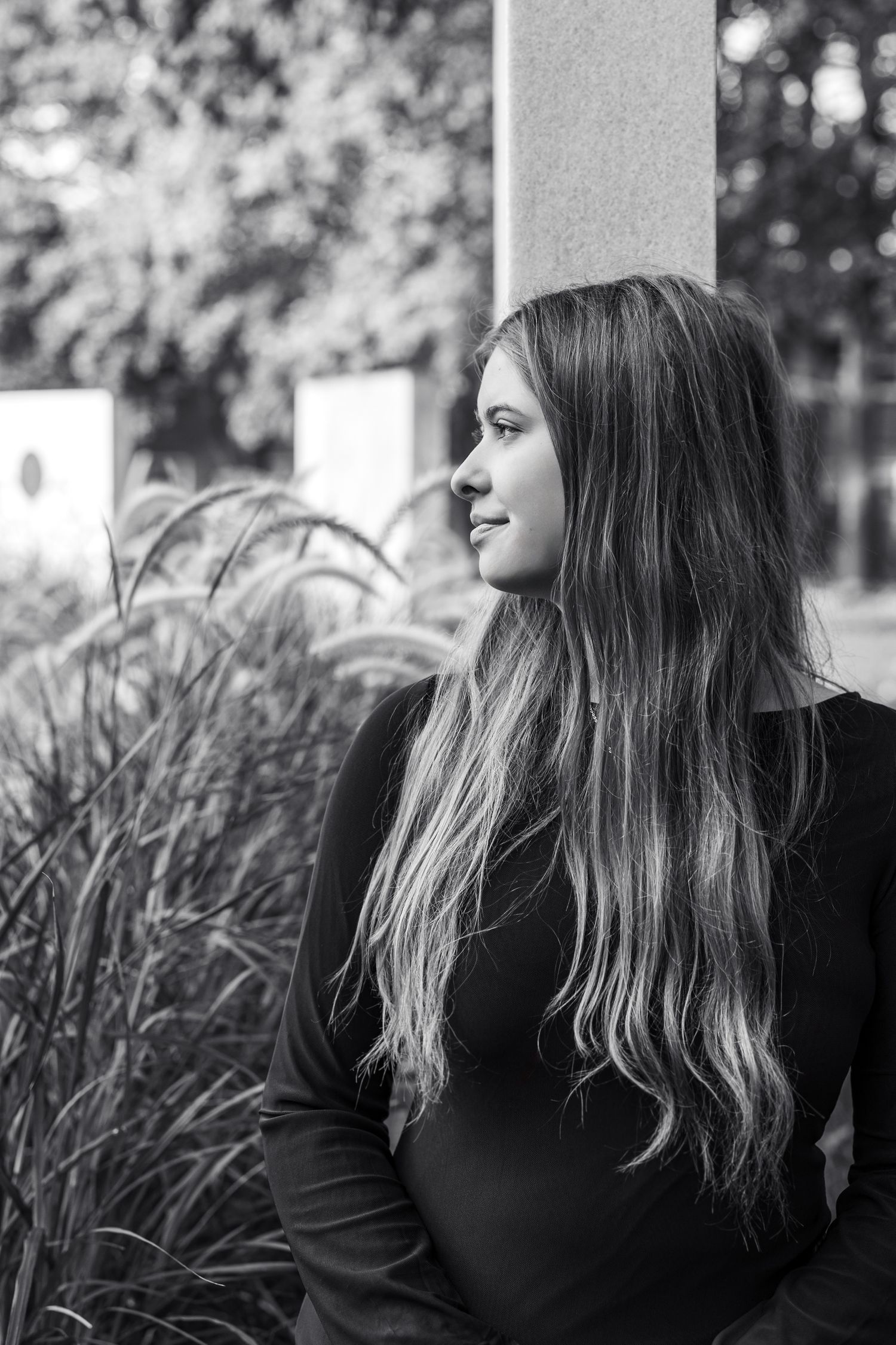 Black and white portrait of a young woman with long wavy hair wearing a black top, gazing thoughtfully outdoors.