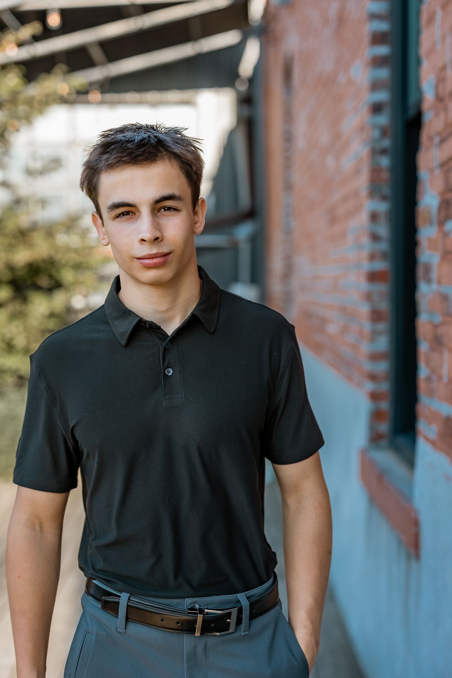 Young man in black polo shirt and gray pants standing outdoors near a brick wall, looking at camera.