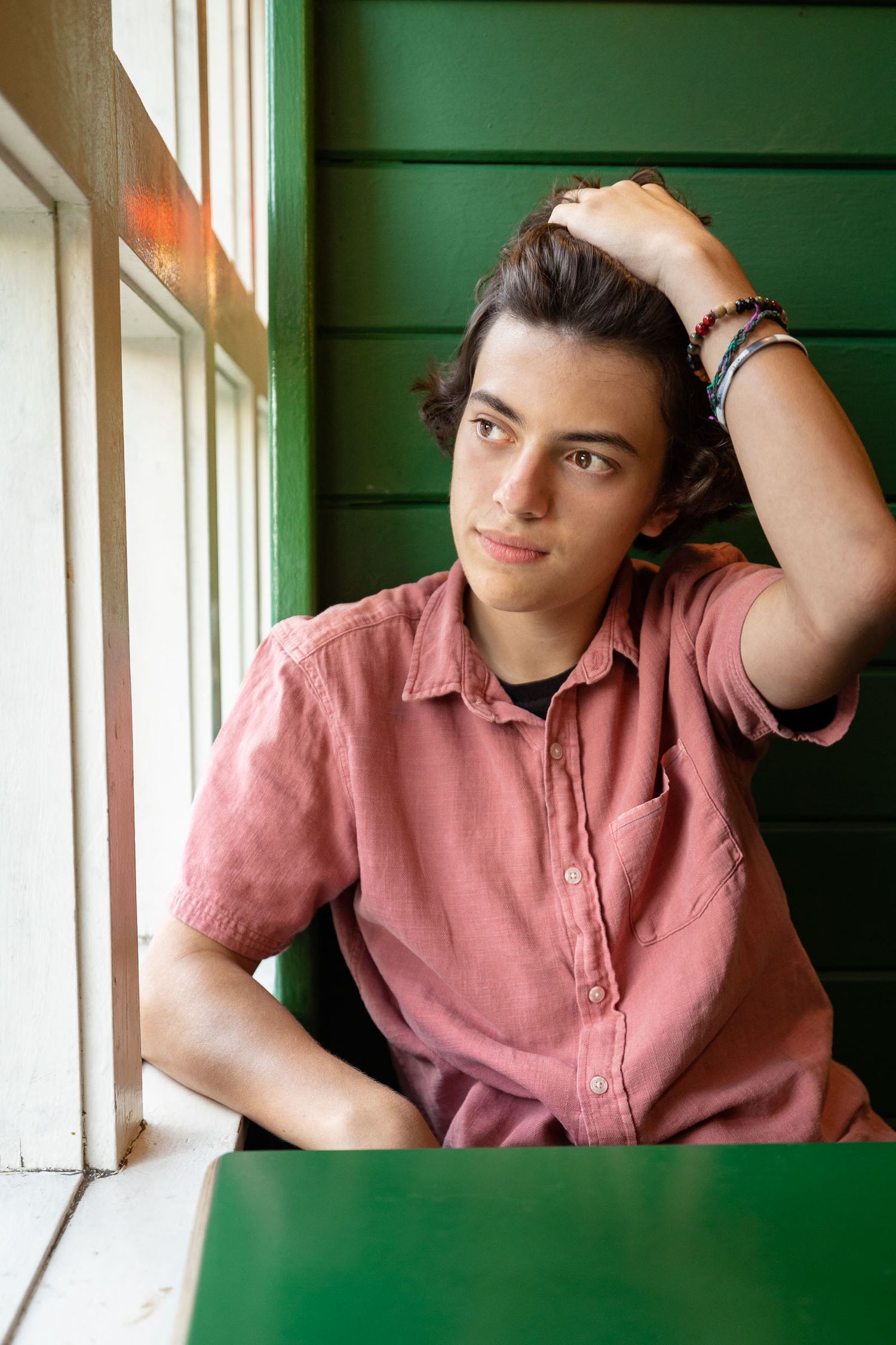 Young man in pink shirt sitting by green wall window, hand in dark hair, wearing bracelets, thoughtful expression.