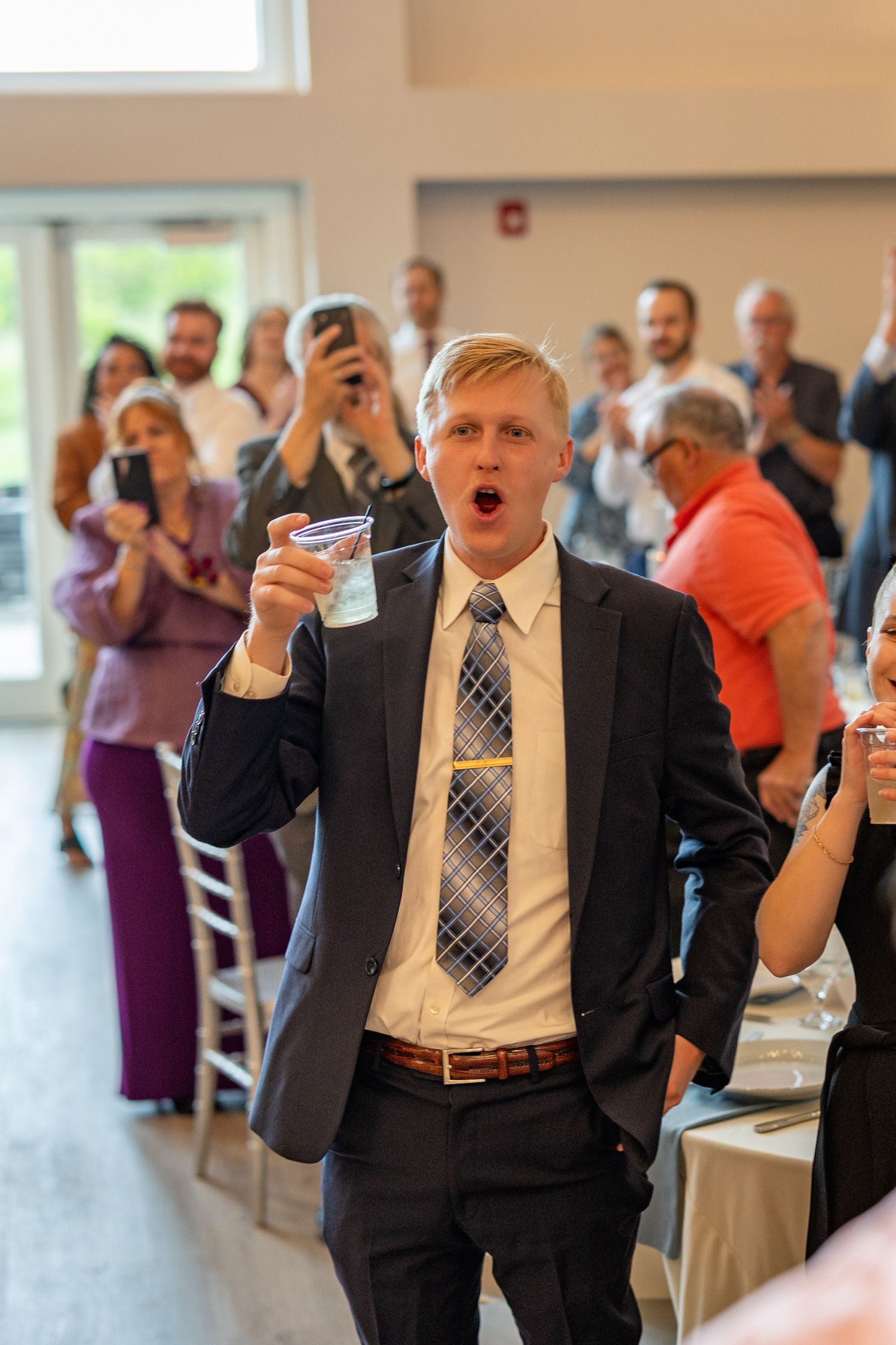 Man in suit raising a glass during a wedding toast while guests applaud and photograph the moment.