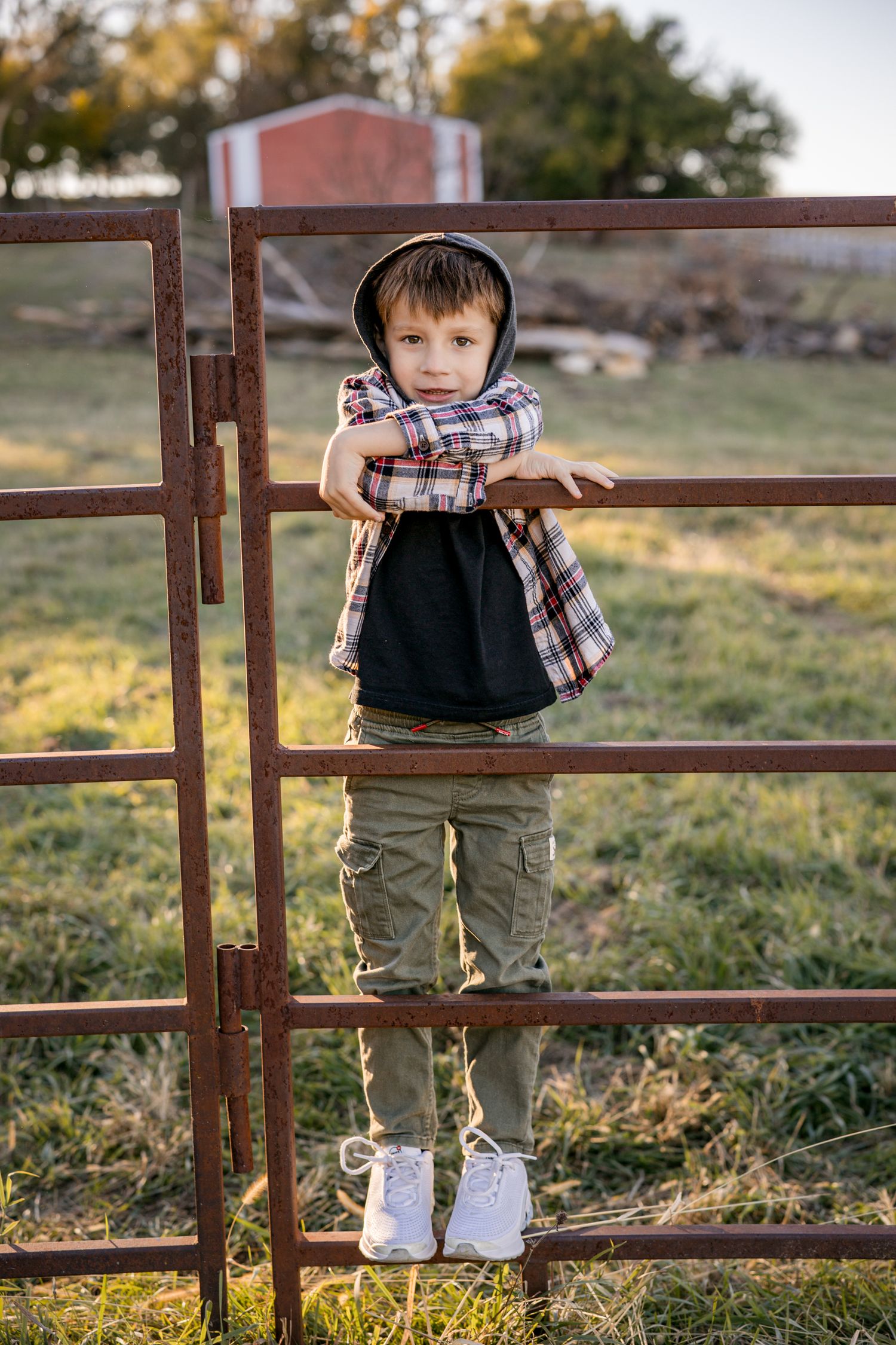 Young boy in plaid shirt and cargo pants leaning on rustic farm gate in golden hour light.