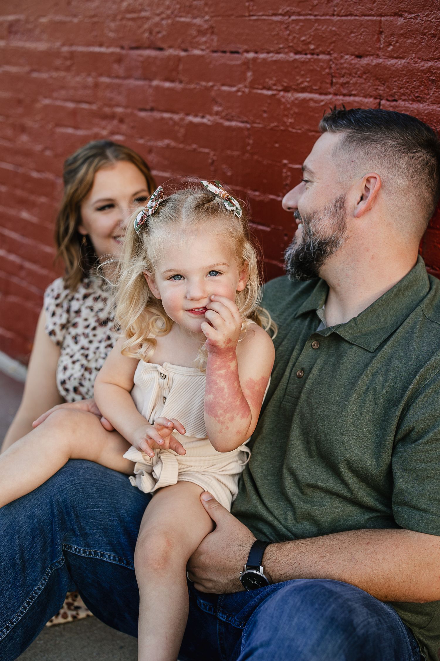 A young family with a blonde toddler girl sits against a red brick wall, smiling and laughing together outdoors.
