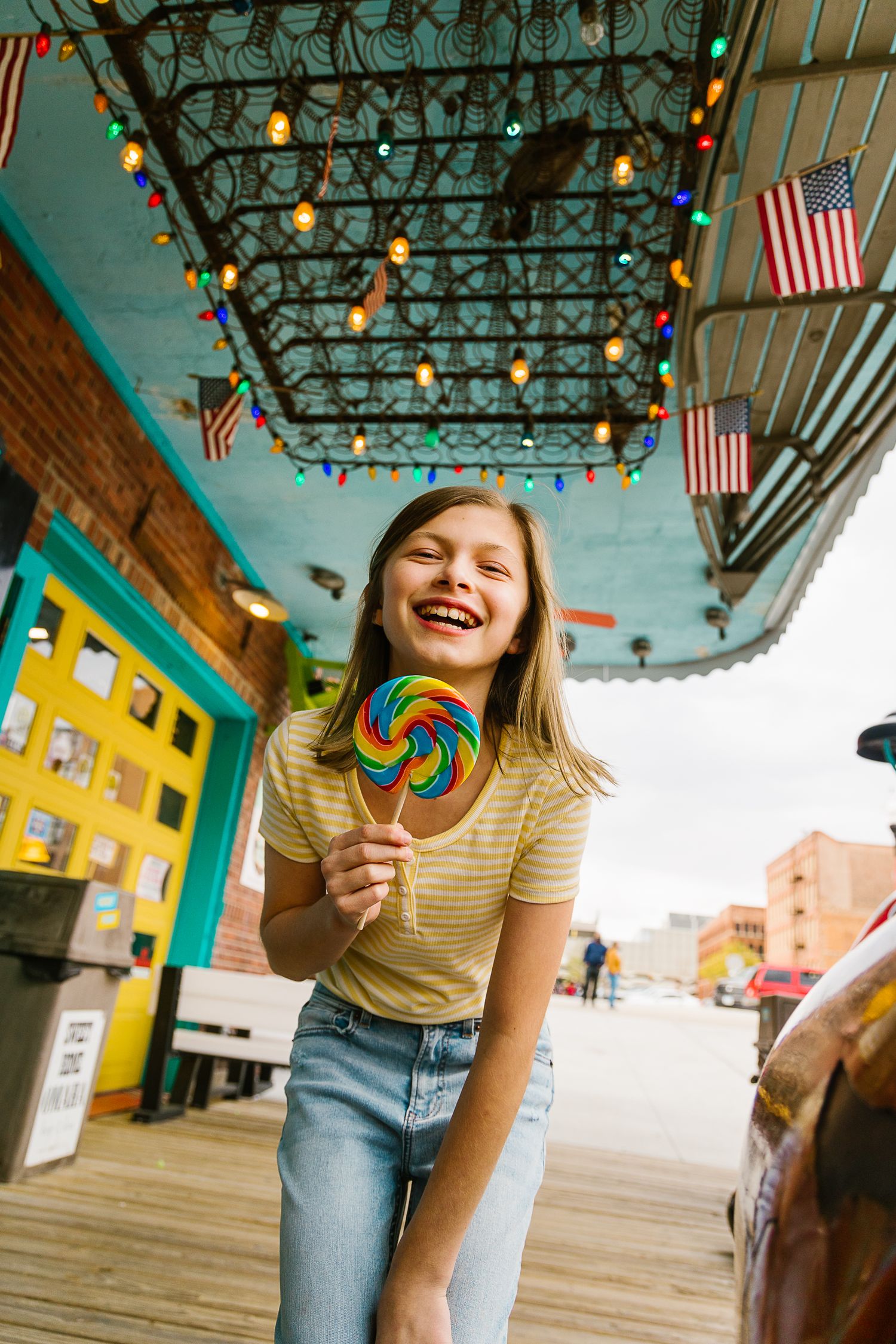 Smiling girl holding colorful rainbow lollipop under string lights at a vibrant American candy shop.