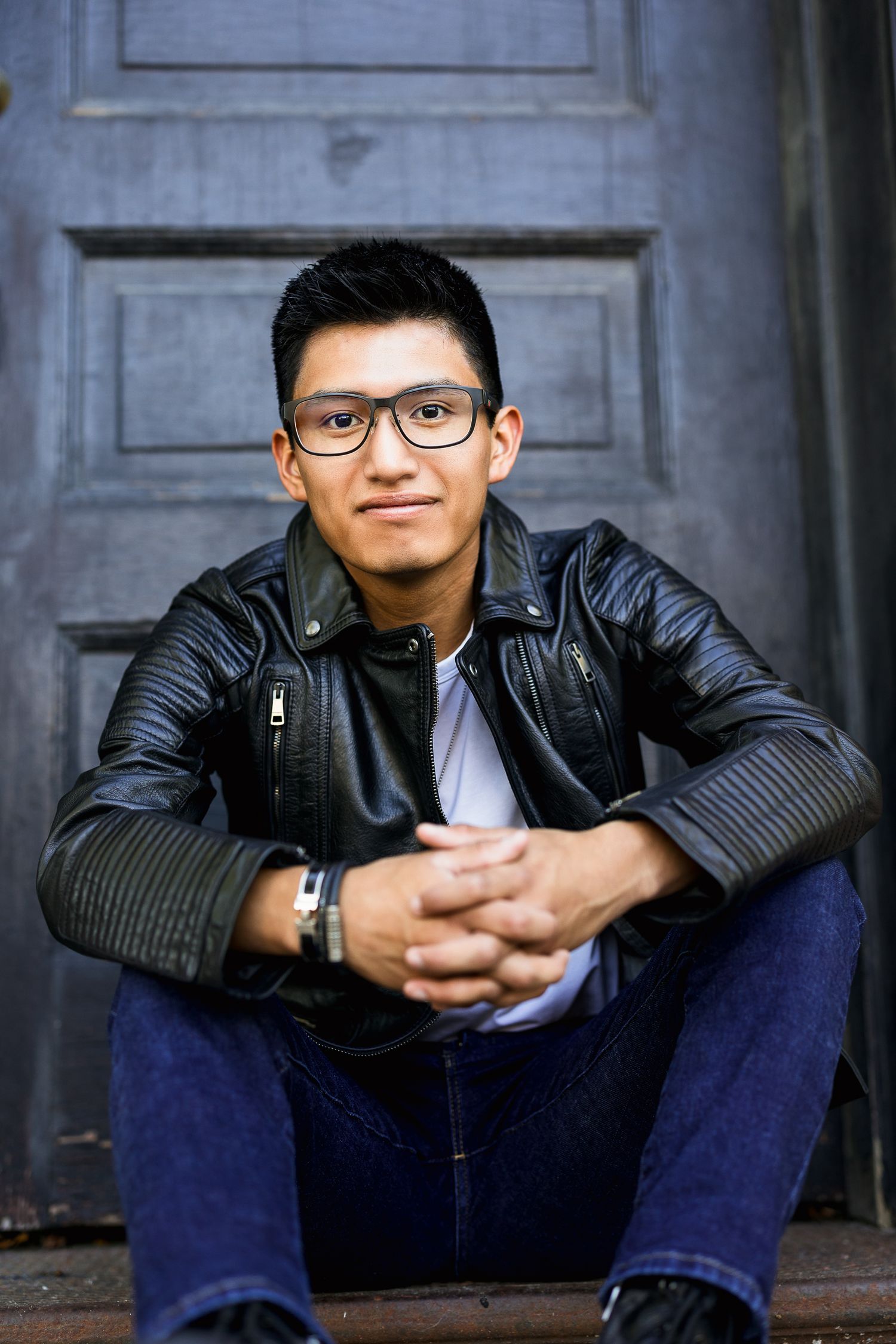 Young man in black leather jacket and glasses sits on steps smiling, wearing jeans and a watch.