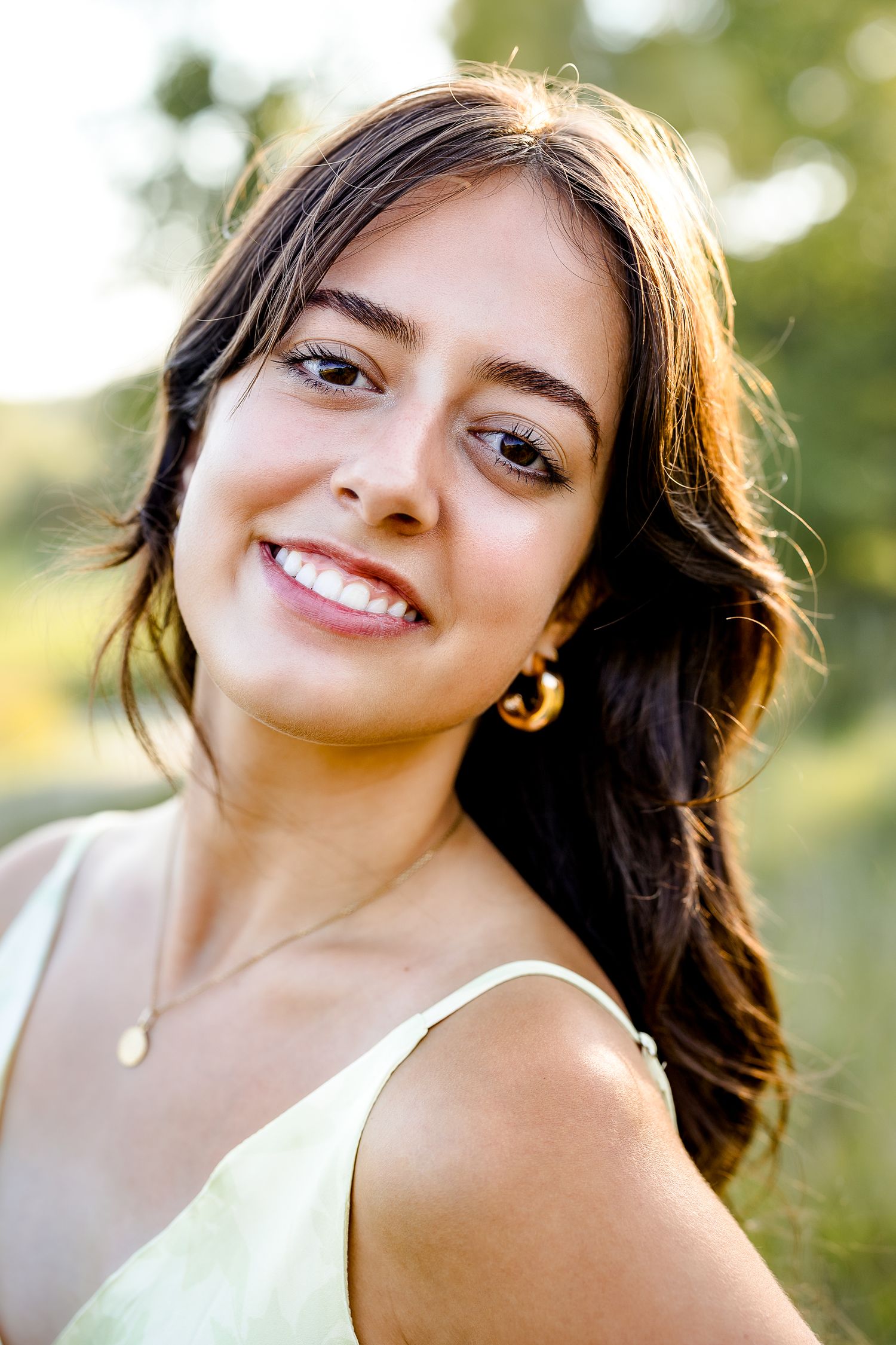 Young woman with long dark hair smiling outdoors, wearing gold hoop earrings and a light green dress.
