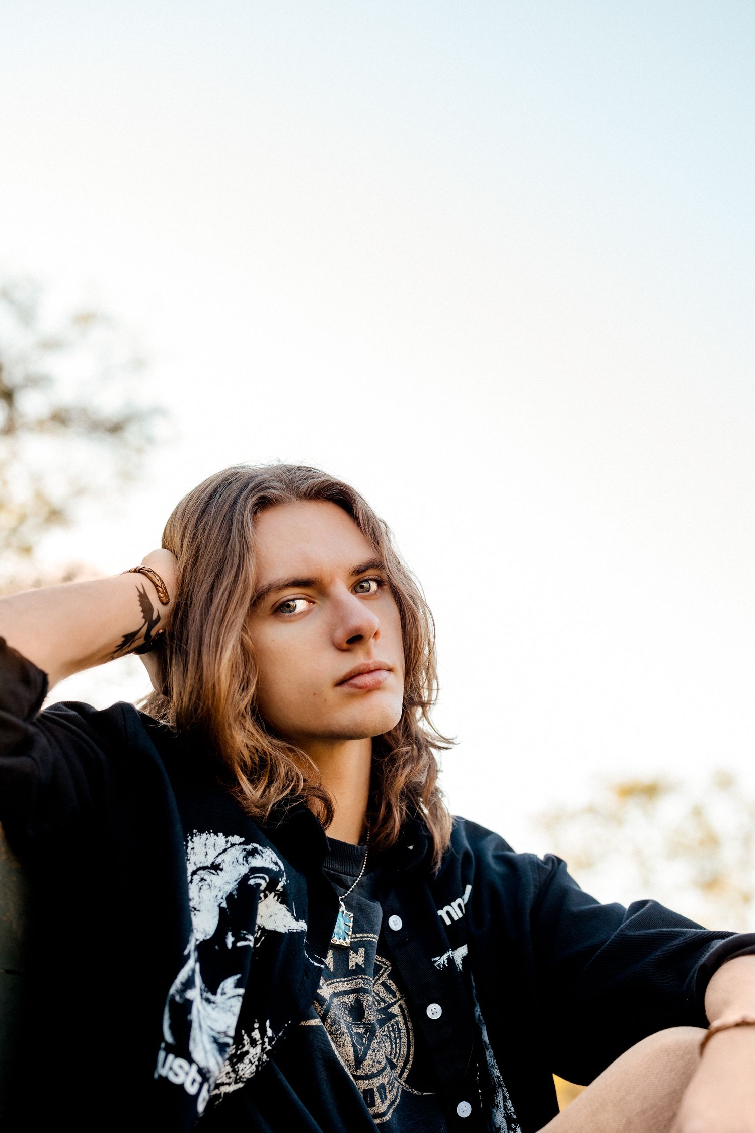 Young man with long hair wearing a black graphic tee sits outdoors against a bright sky backdrop.