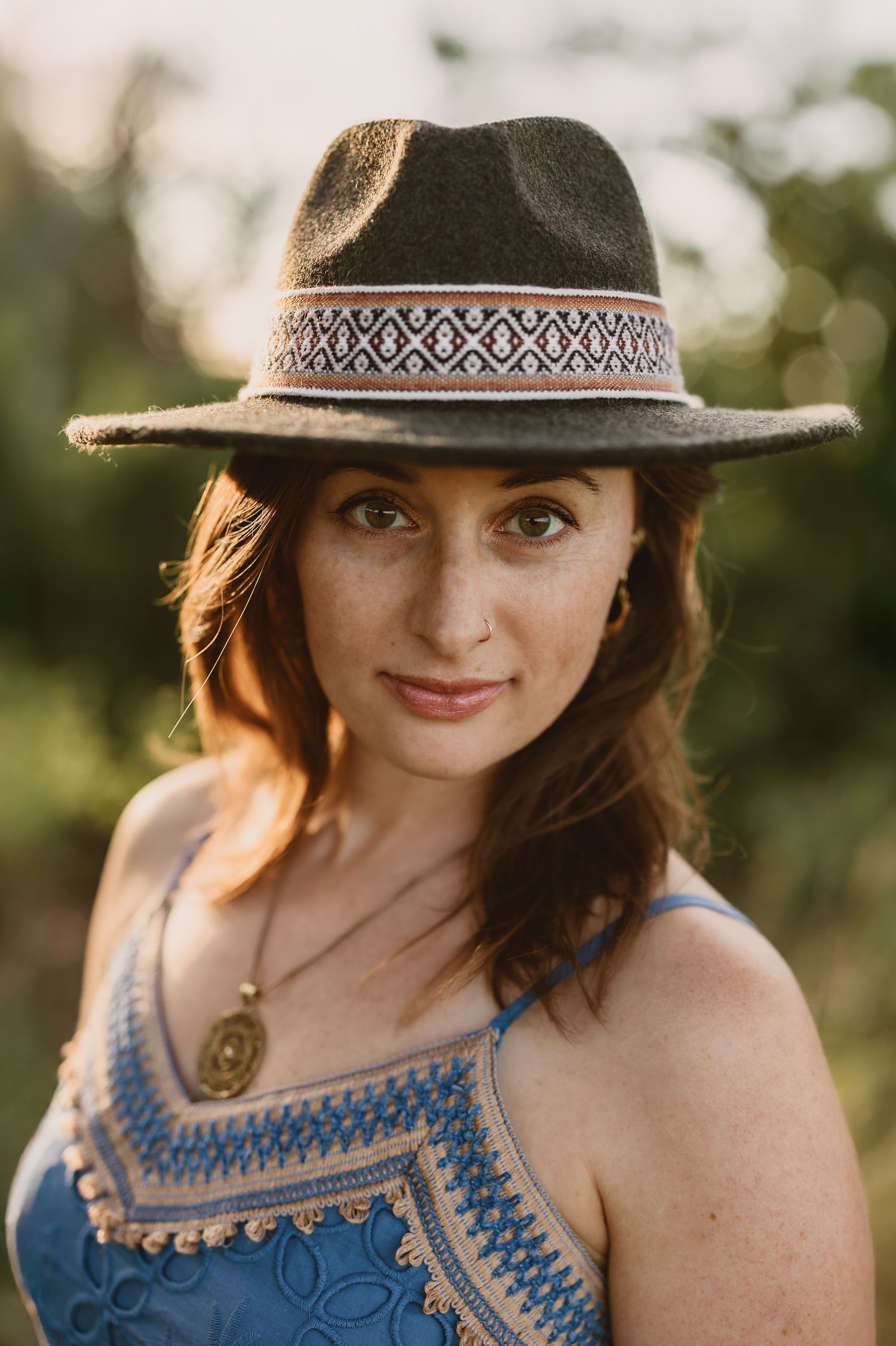 A person in a brown hat and blue patterned tank top smiles while posing outdoors during golden hour.
