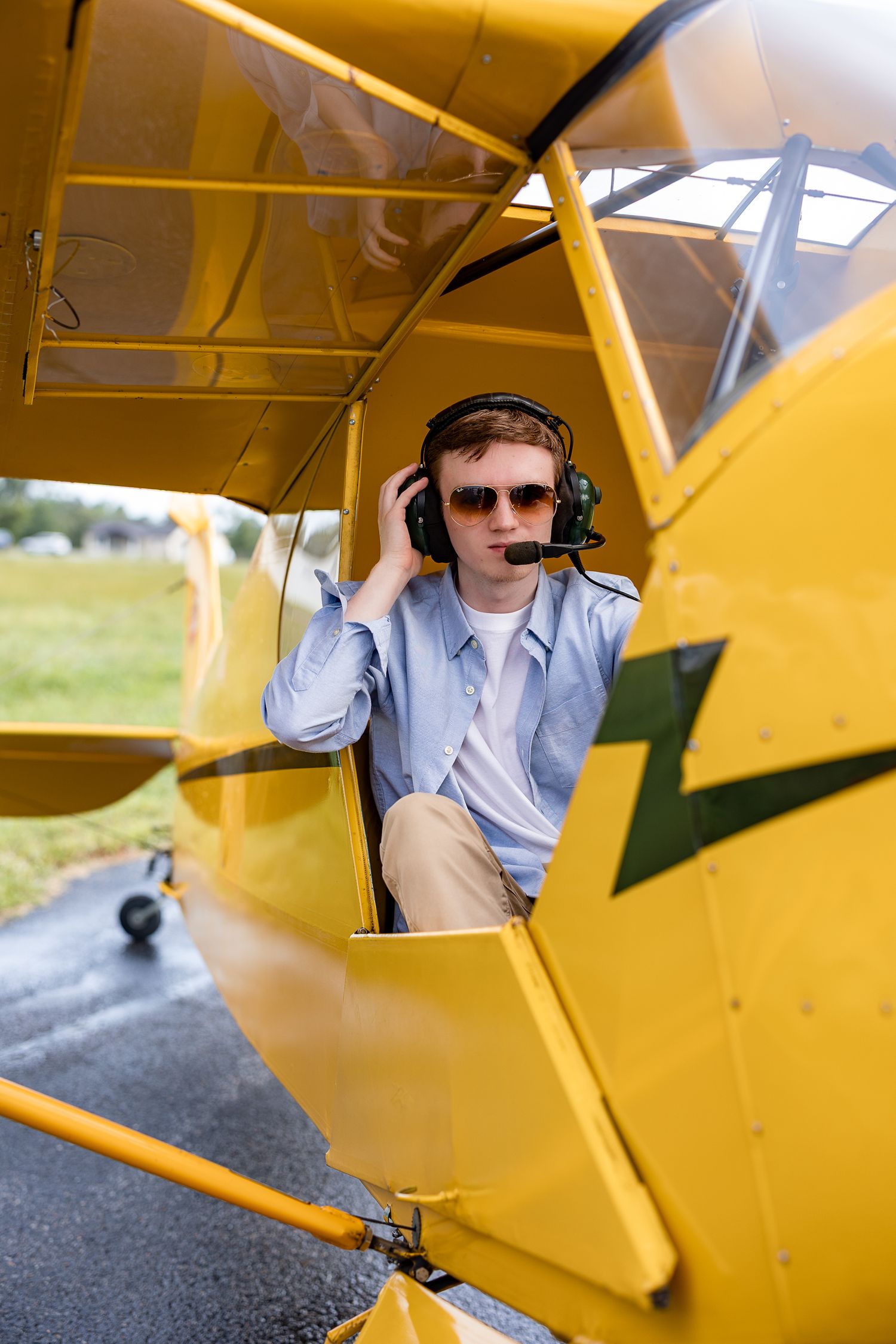 Young pilot in sunglasses and headset sits in yellow small aircraft cockpit on airstrip.