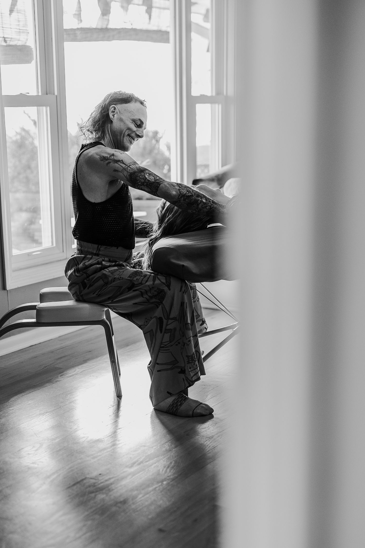 Black and white photo of a tattooed person sitting on a chair, smiling near a bright window indoors.