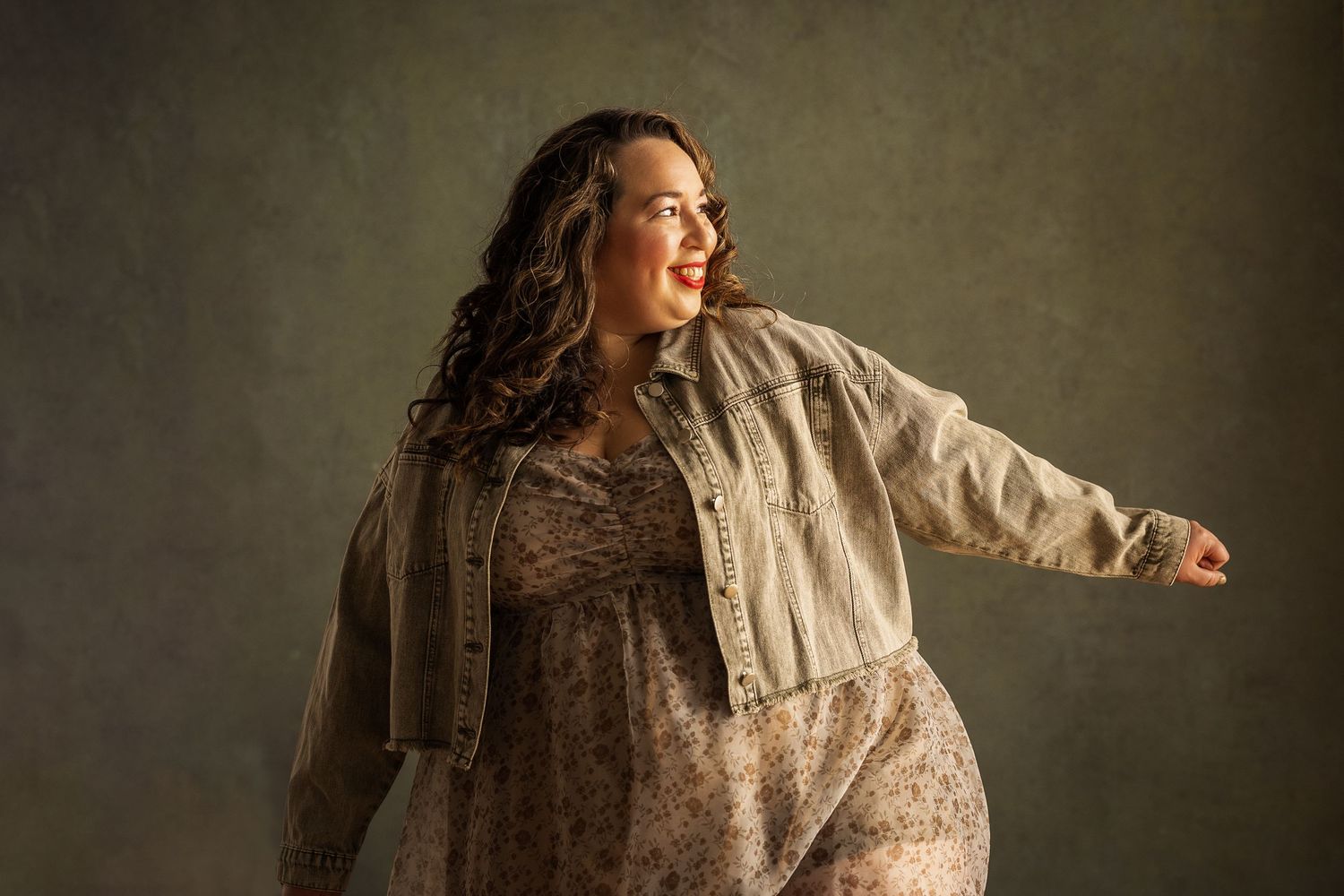A woman in a floral dress and denim jacket poses gracefully against a moody olive-green backdrop.