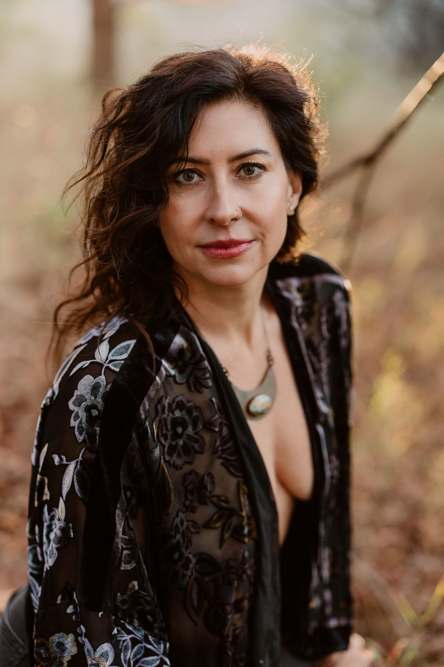 Curly-haired woman in floral black velvet jacket with necklace smiling outdoors in autumn forest.