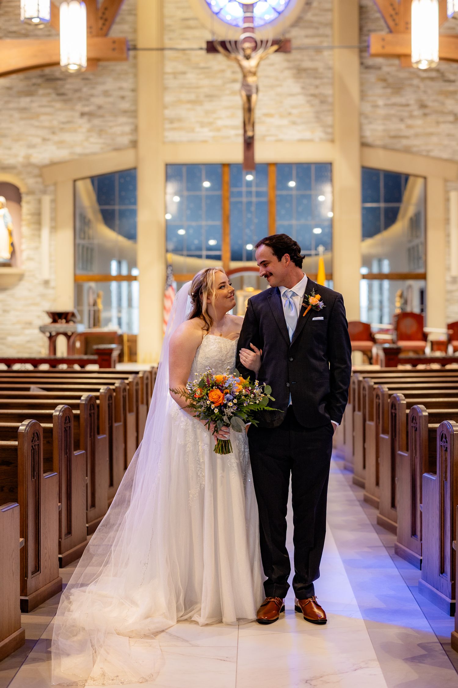 Bride and groom walk down church aisle, smiling at each other, bride holding colorful bouquet, crucifix visible in background.