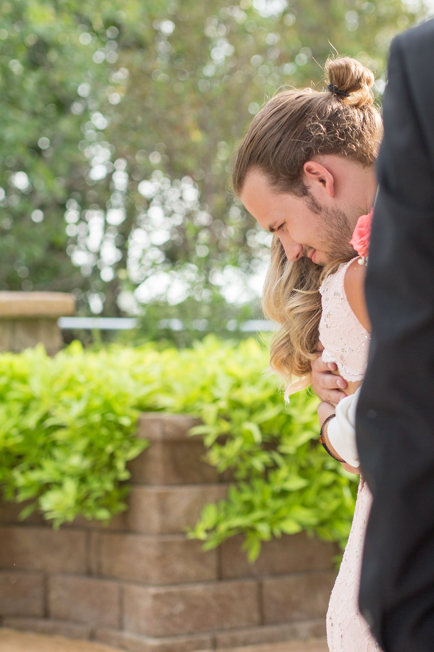 Groom with man bun tenderly embracing blonde bride in white lace dress in sunlit garden setting.