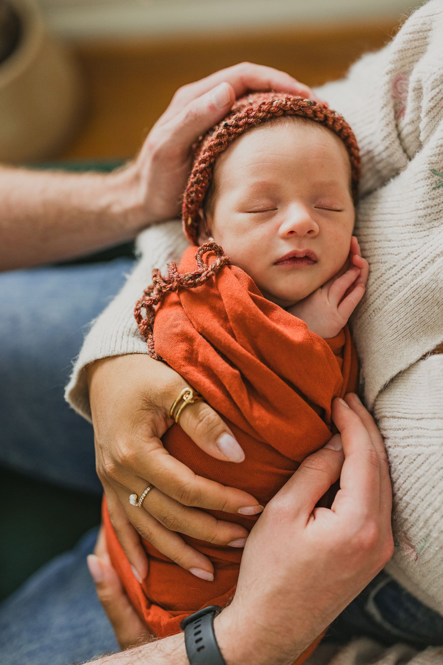 Newborn baby sleeping peacefully in parents arms, wrapped in orange swaddle with matching knit bonnet.