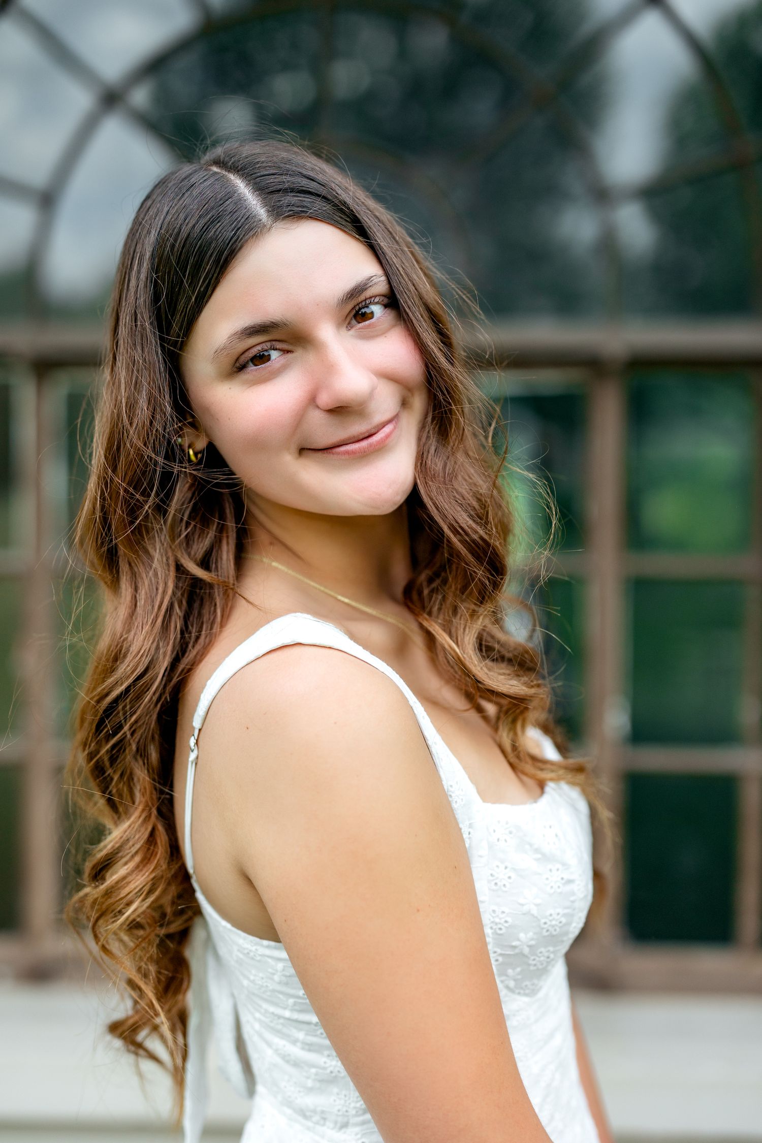 Young woman with long wavy brown hair smiles over shoulder in white dress near greenhouse windows.