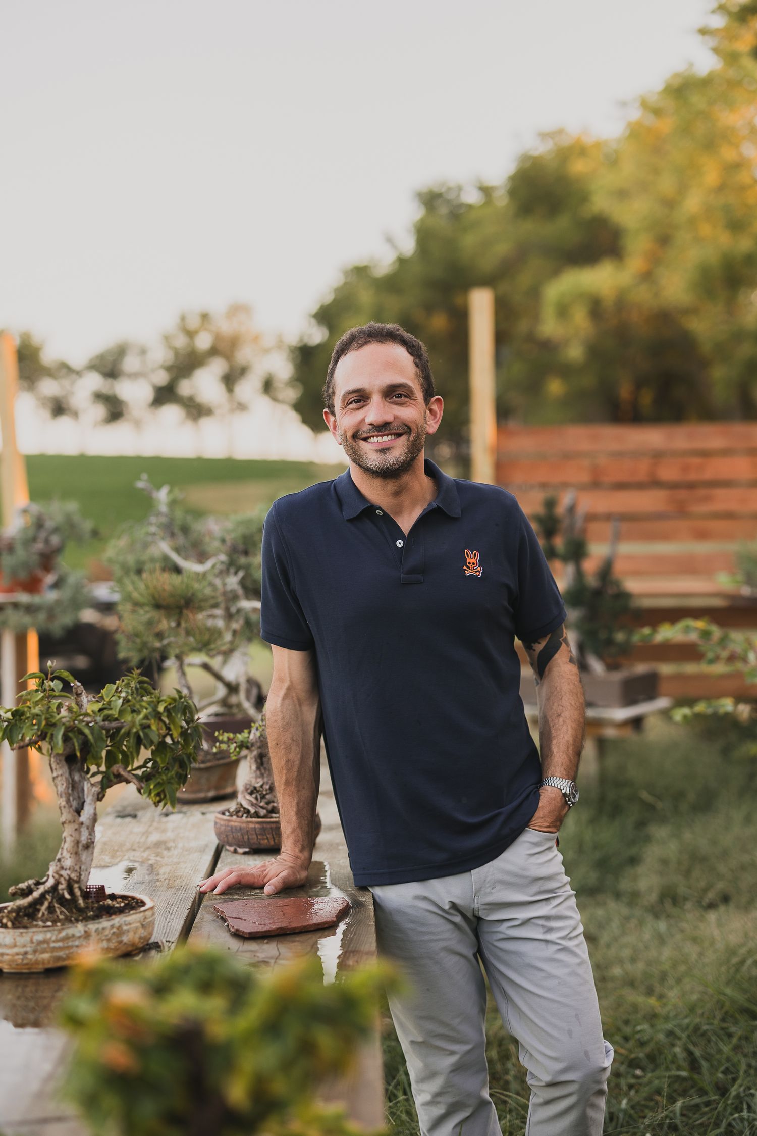 A small business owner in a navy polo shirt and grey pants smiles while standing in a garden setting at sunset.