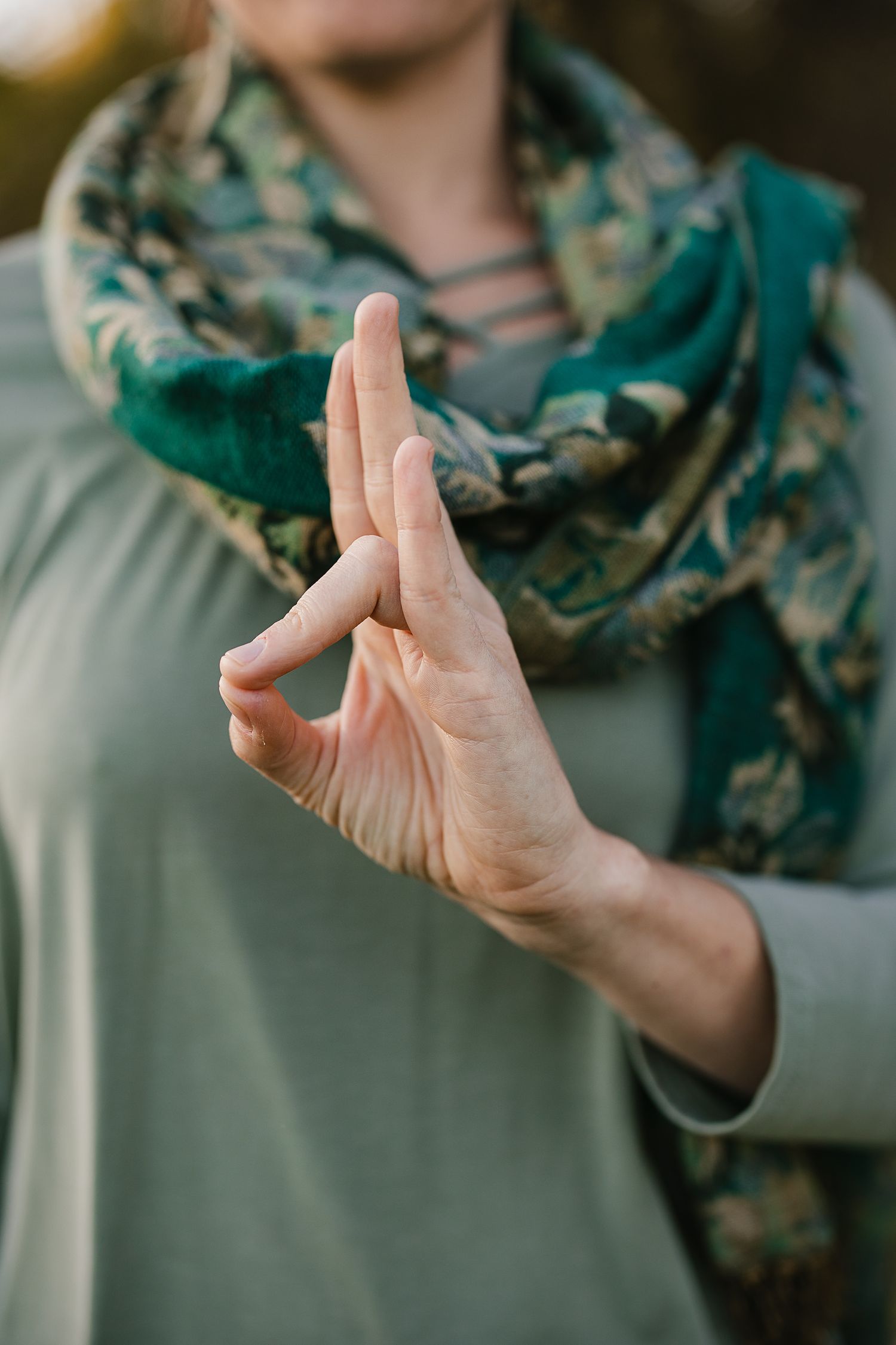Woman in sage green dress performing a mudra hand gesture, wearing a floral teal scarf, practicing yoga meditation outdoors.