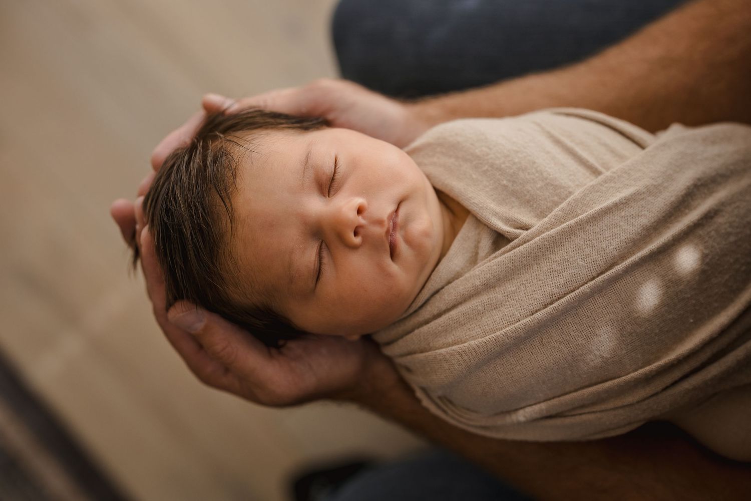 Sleeping newborn baby swaddled in a beige wrap, cradled gently in adult hands during a newborn photography session.