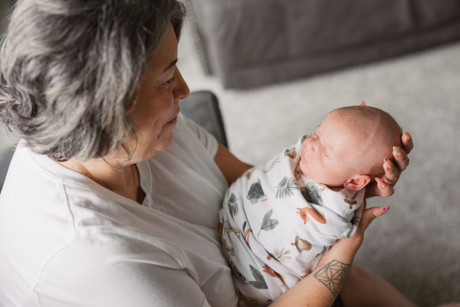 Grandmother with gray hair smiling while holding a sleeping newborn baby wrapped in a white floral swaddle blanket.