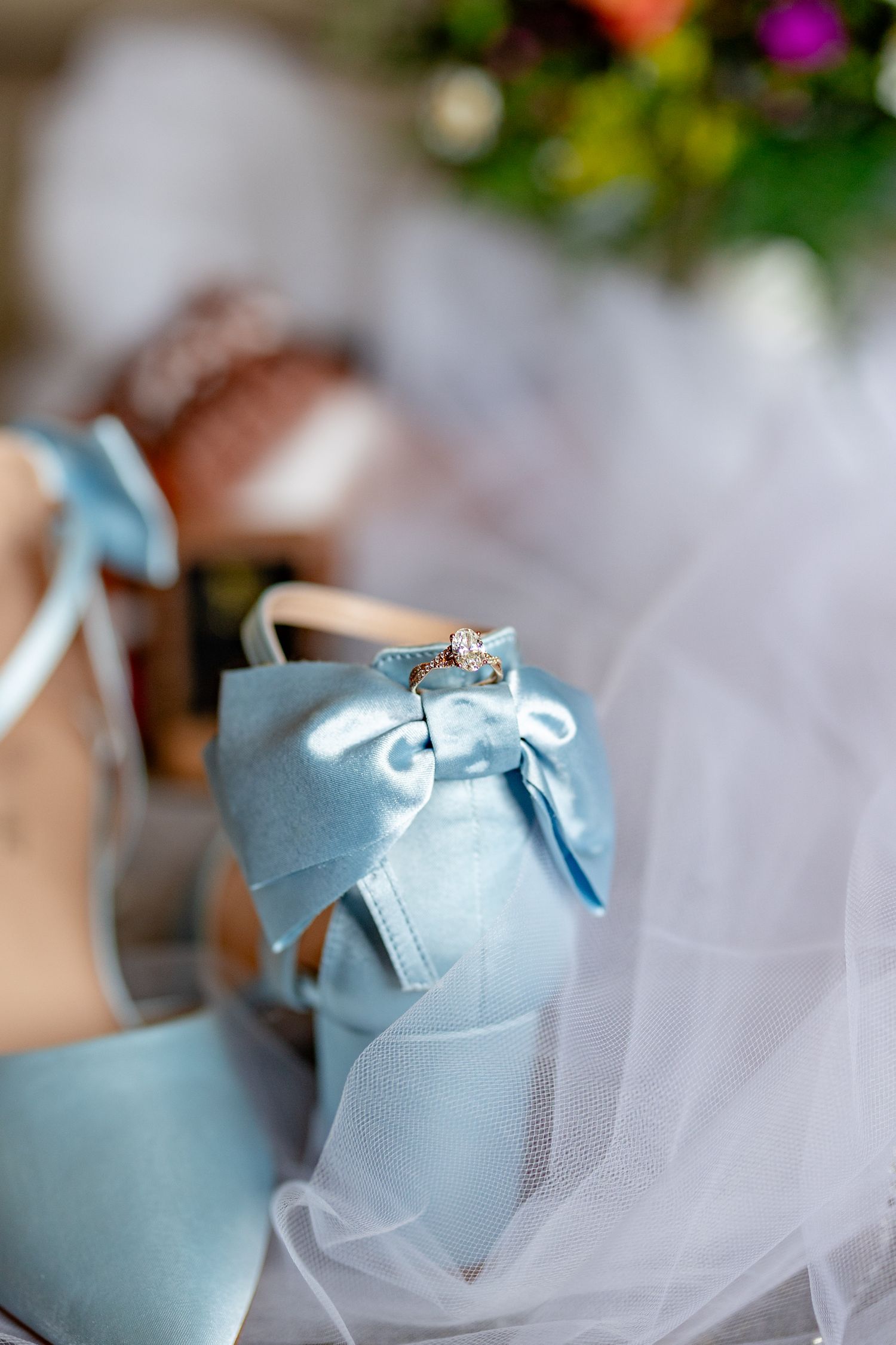 Light blue satin bridal heels with bow detail and diamond ring, resting on white veil with colorful bouquet in background.