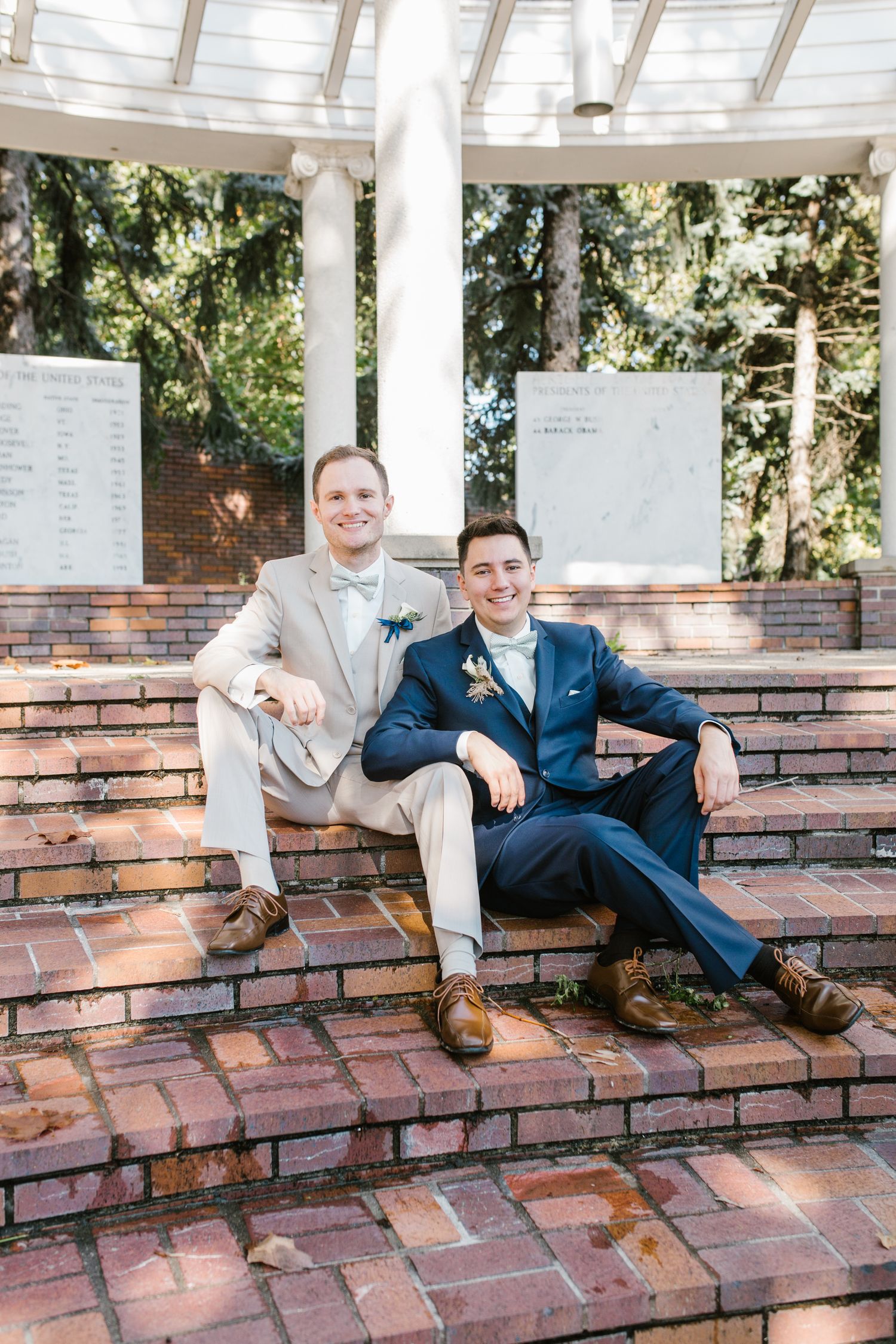 Two grooms in suits sitting on brick steps at an outdoor wedding venue, smiling for a portrait photo.