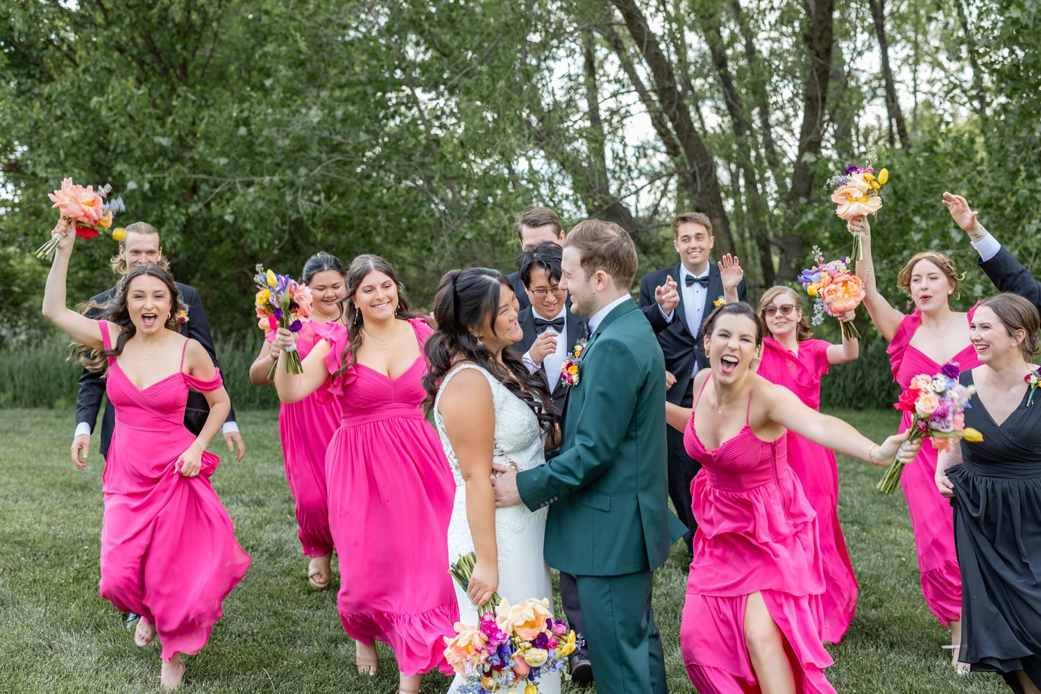 Wedding party celebrates bride and groom outdoors, bridesmaids in vibrant pink dresses cheering among lush green trees.