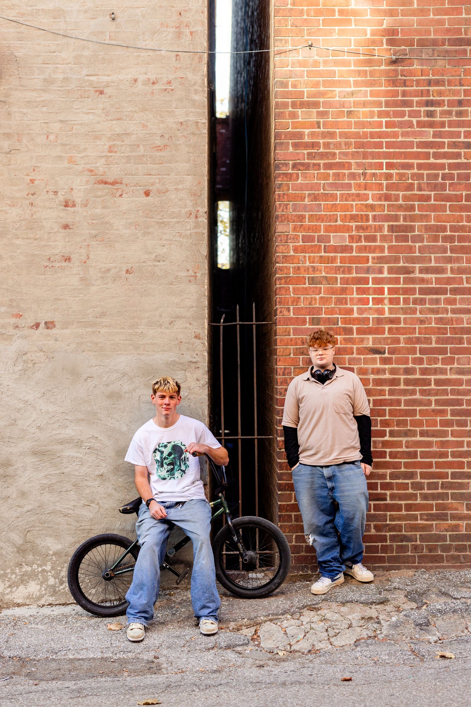 Two teenage boys posing near brick wall alley, one sitting on BMX bike wearing white tee, other leaning against wall in tan shirt.