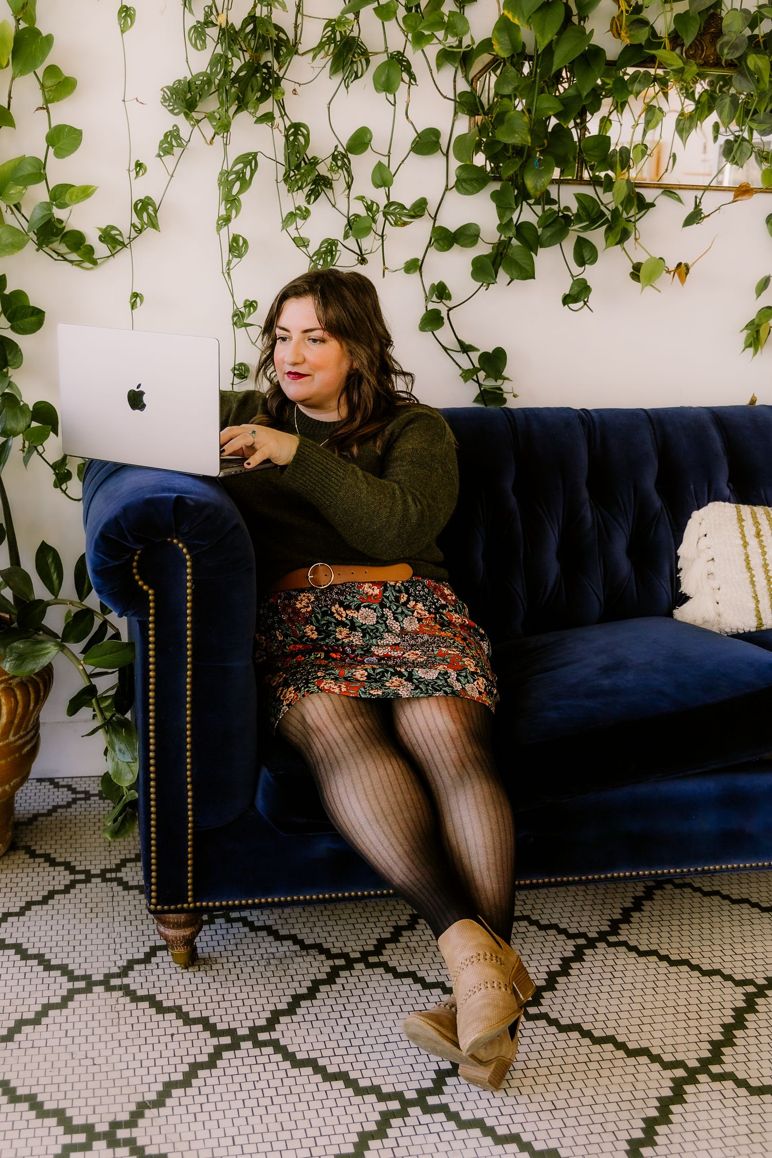 Woman in floral skirt and green sweater working on MacBook on blue velvet sofa surrounded by lush indoor plants.