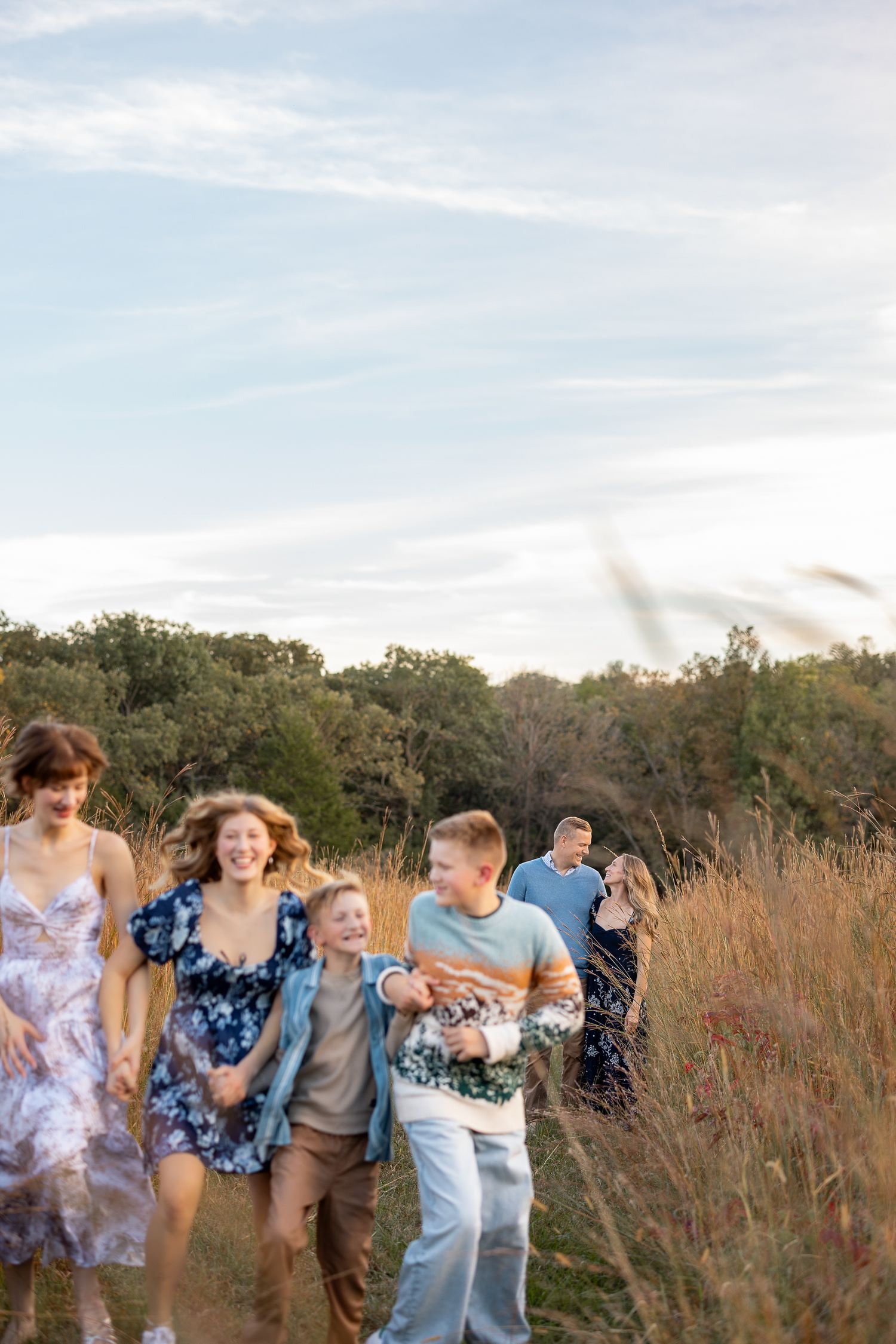 A joyful family runs through a golden field with trees and blue sky, laughing together during an outdoor photo session.
