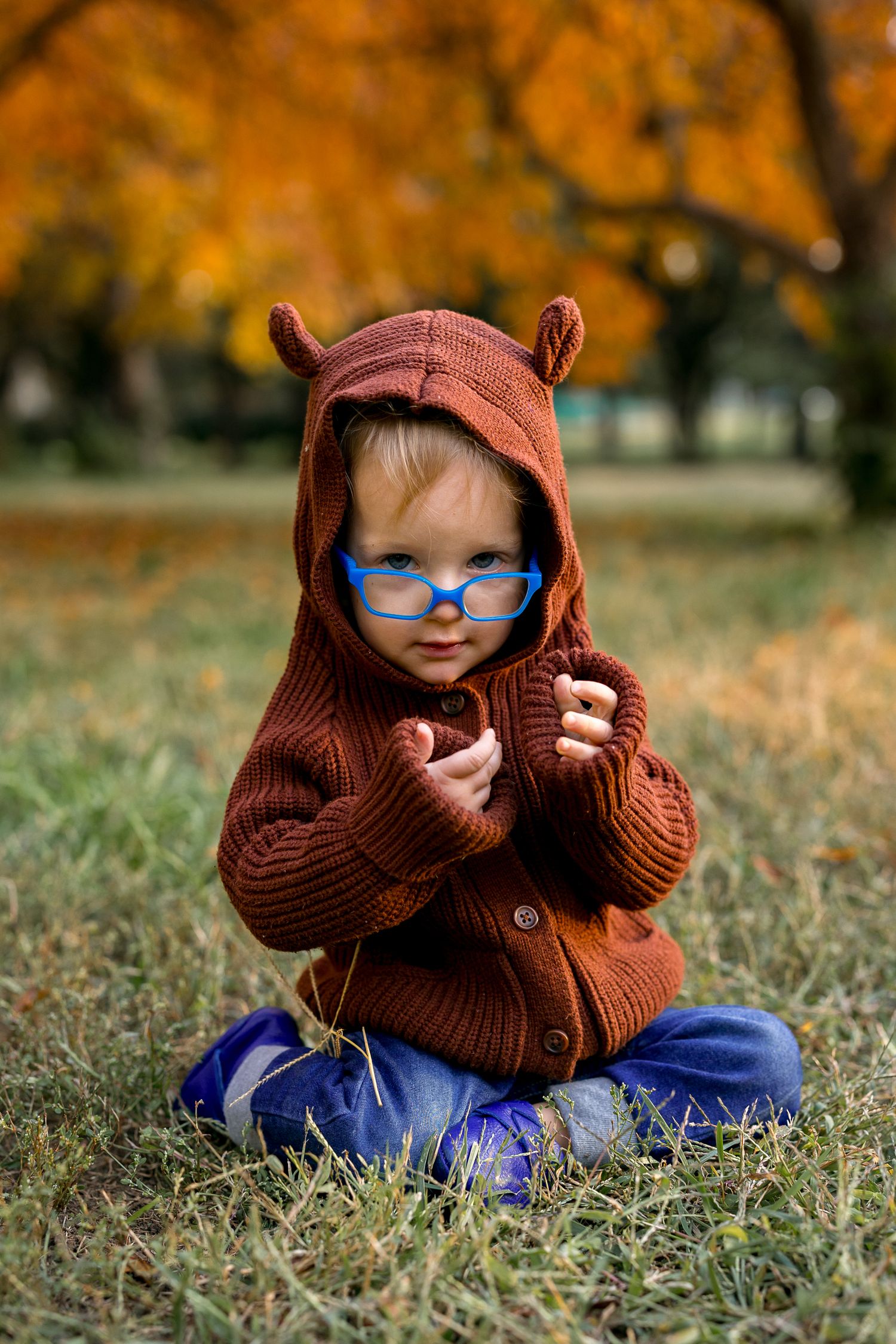 Toddler in brown bear-eared hoodie and blue glasses sitting cross-legged on autumn grass in a park.