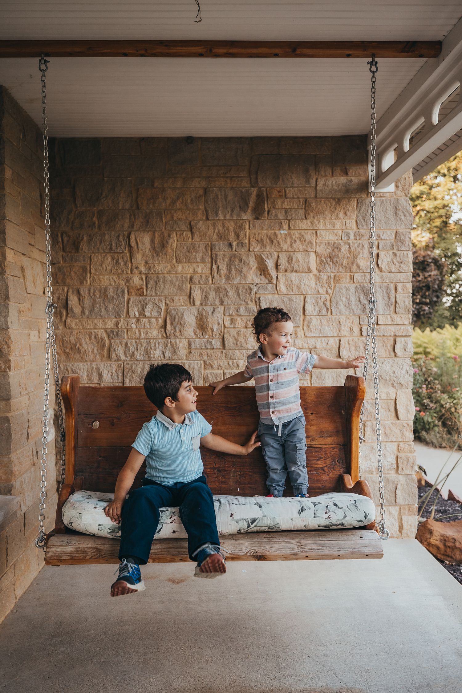 Two young boys playing on a rustic wooden porch swing against a stone wall exterior.