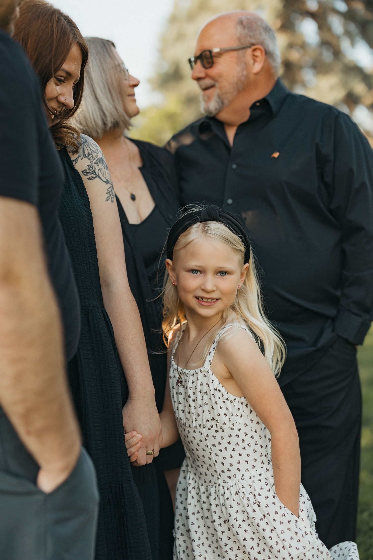 Young blonde girl in floral dress smiles holding hands with family during outdoor photo session.