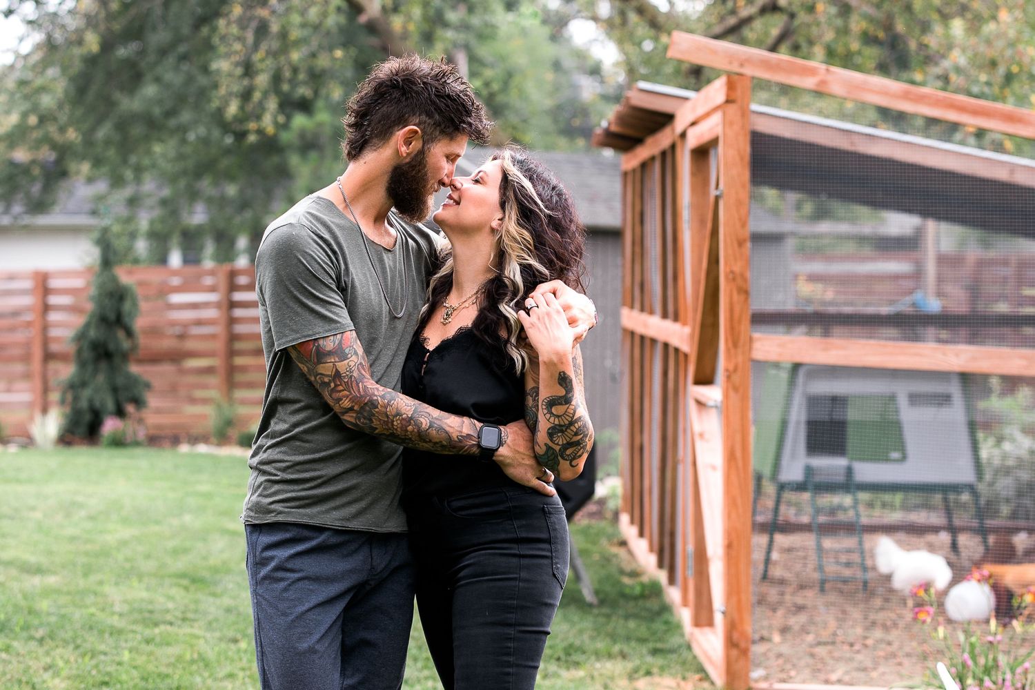 Tattooed couple sharing a romantic moment outdoors near a wooden fence in a lush backyard garden.