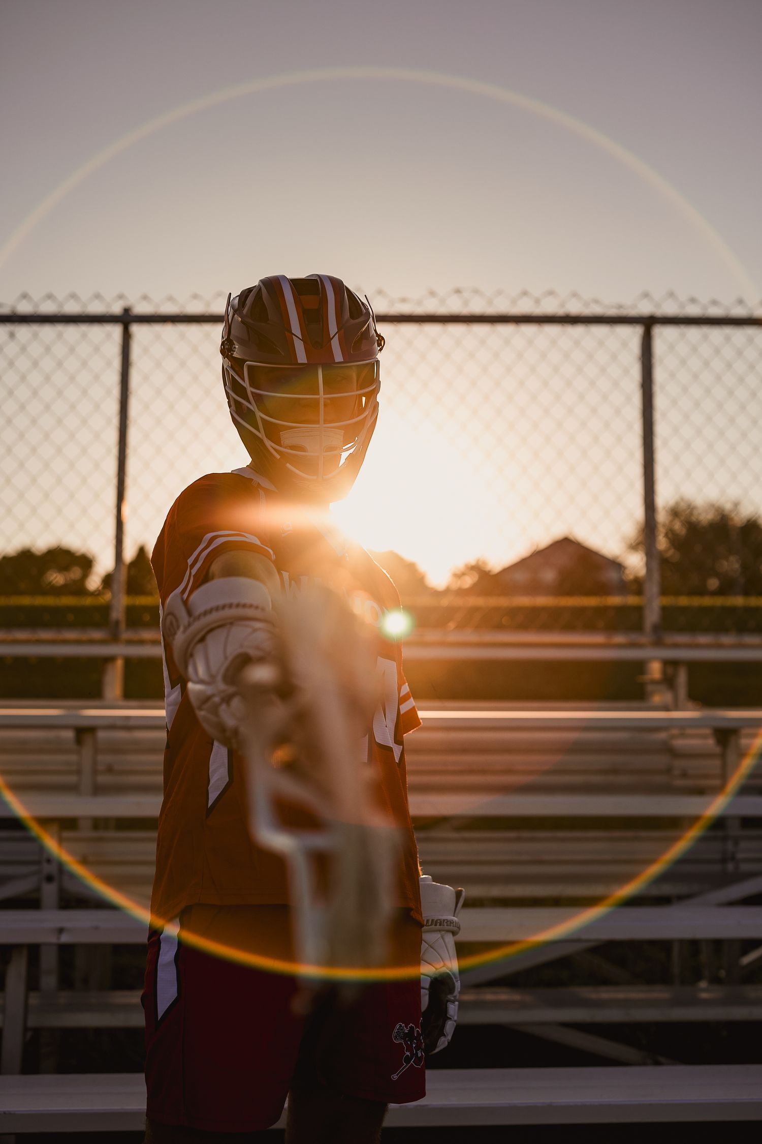 Lacrosse player in helmet and gear holds stick toward camera at sunset with golden lens flare on bleachers.