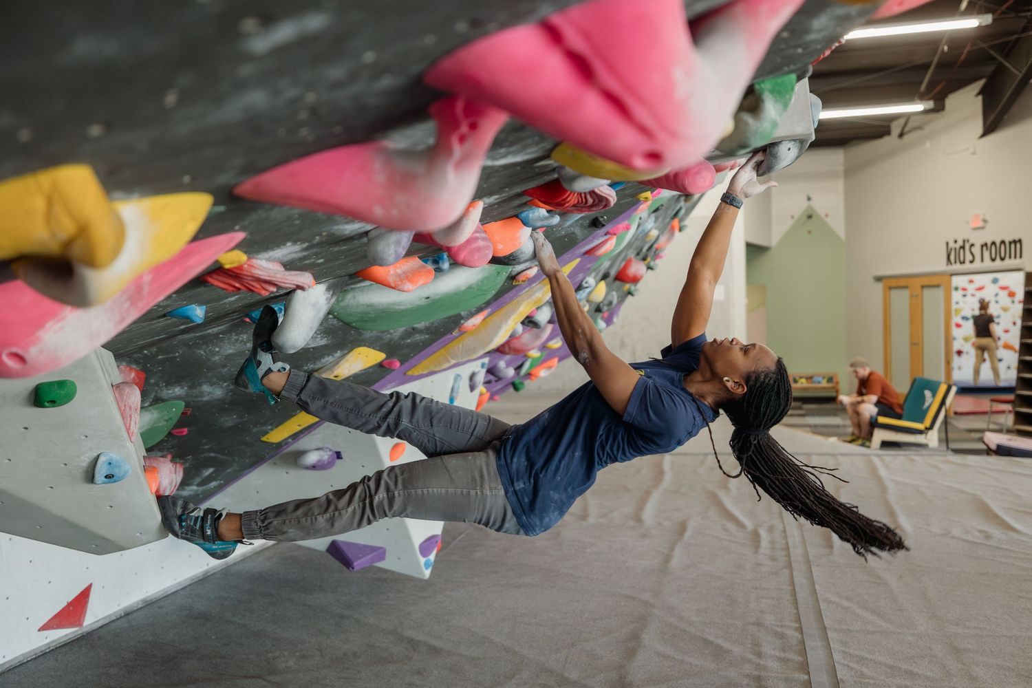 A woman with a long pony tail bouldering on an indoor climbing wall with colorful holds in a gym with a kids room visible in the background.