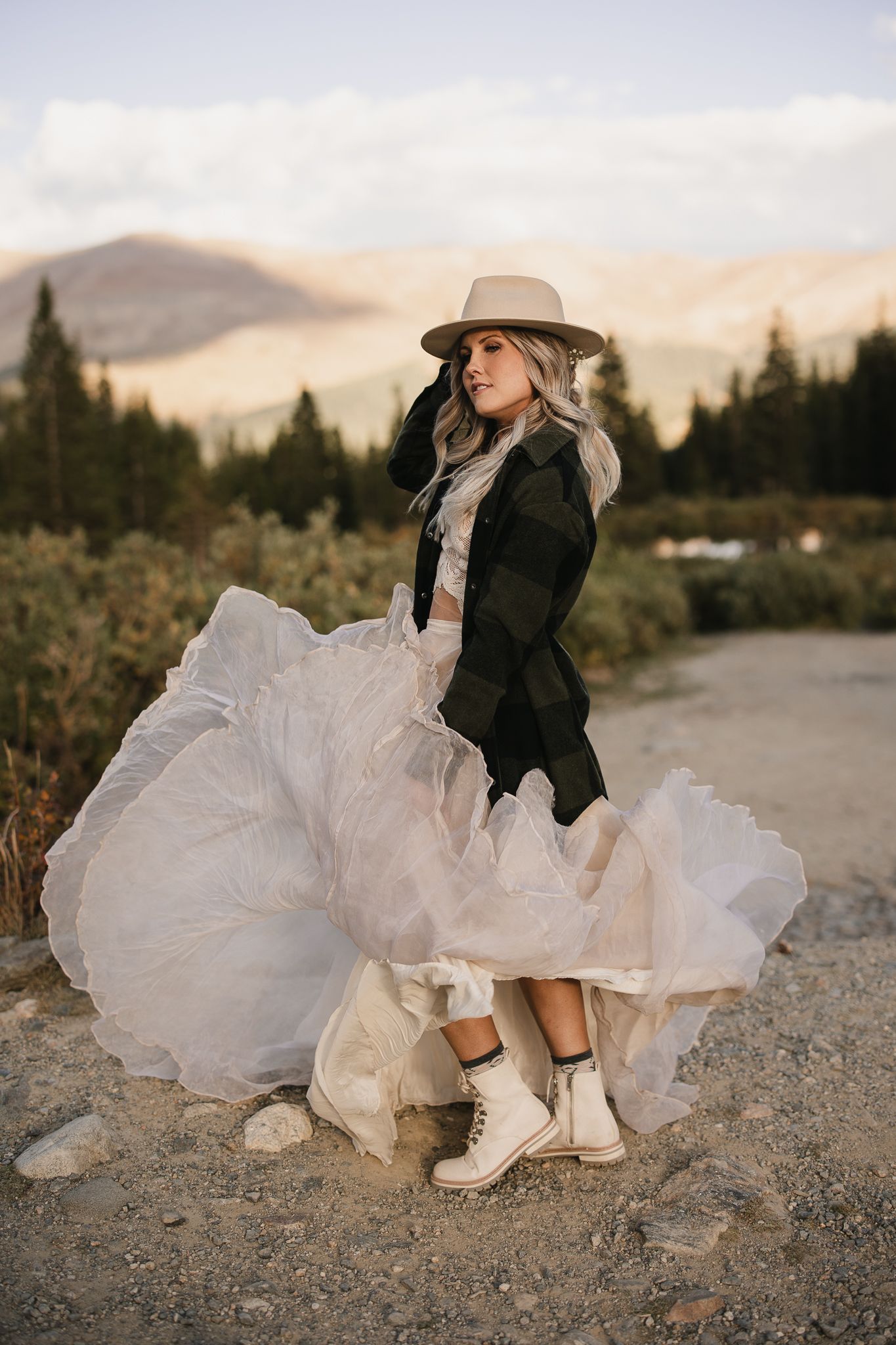 Fashion shoot featuring flowing white dress and cowboy boots against mountain landscape at sunset.