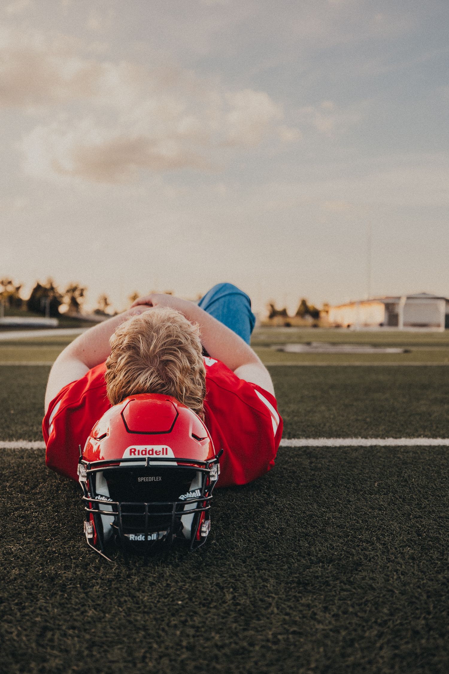 Young football player in red jersey lying on field with Riddell helmet at sunset.