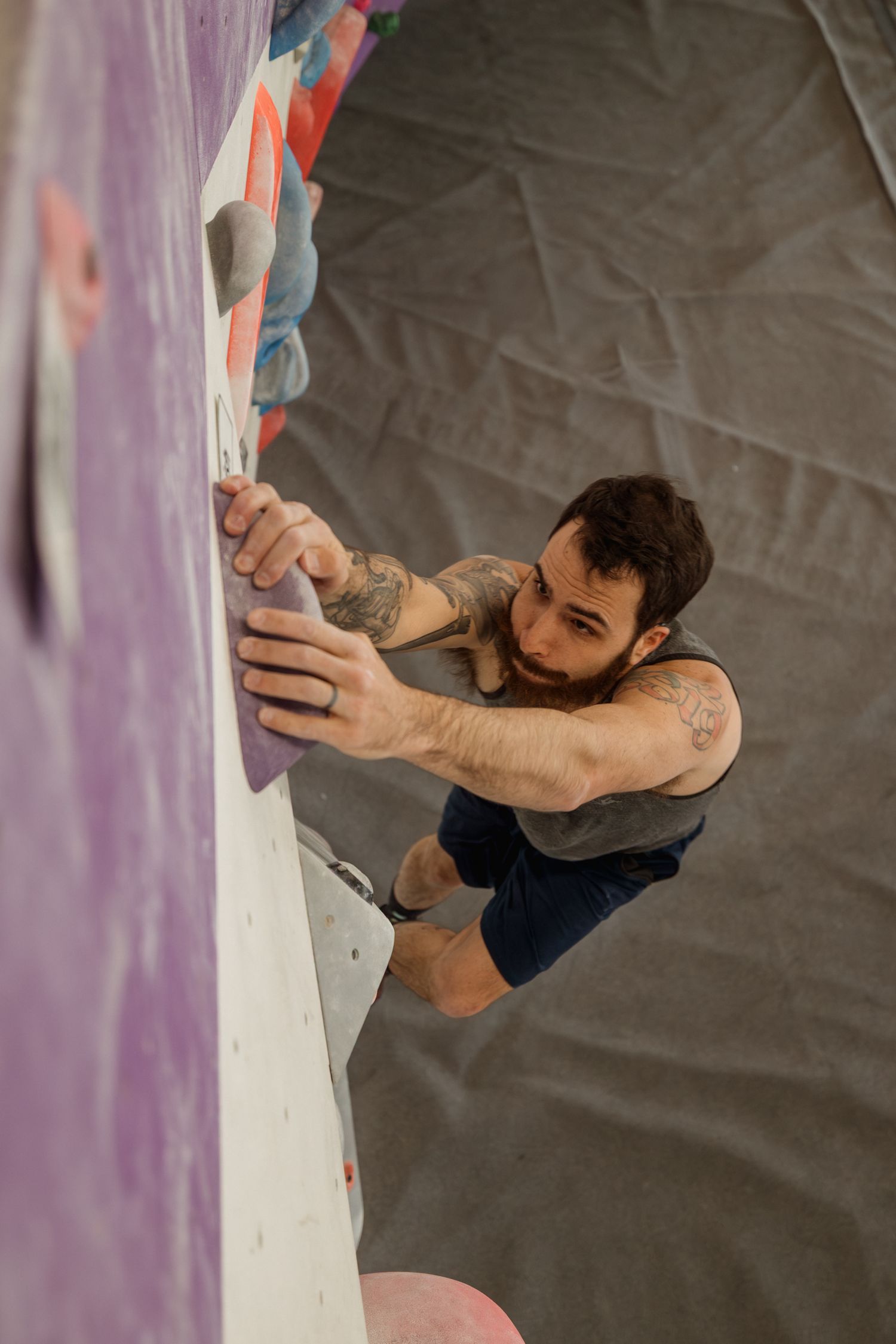 Tattooed man with beard bouldering on indoor climbing wall, gripping colorful holds in a gym.