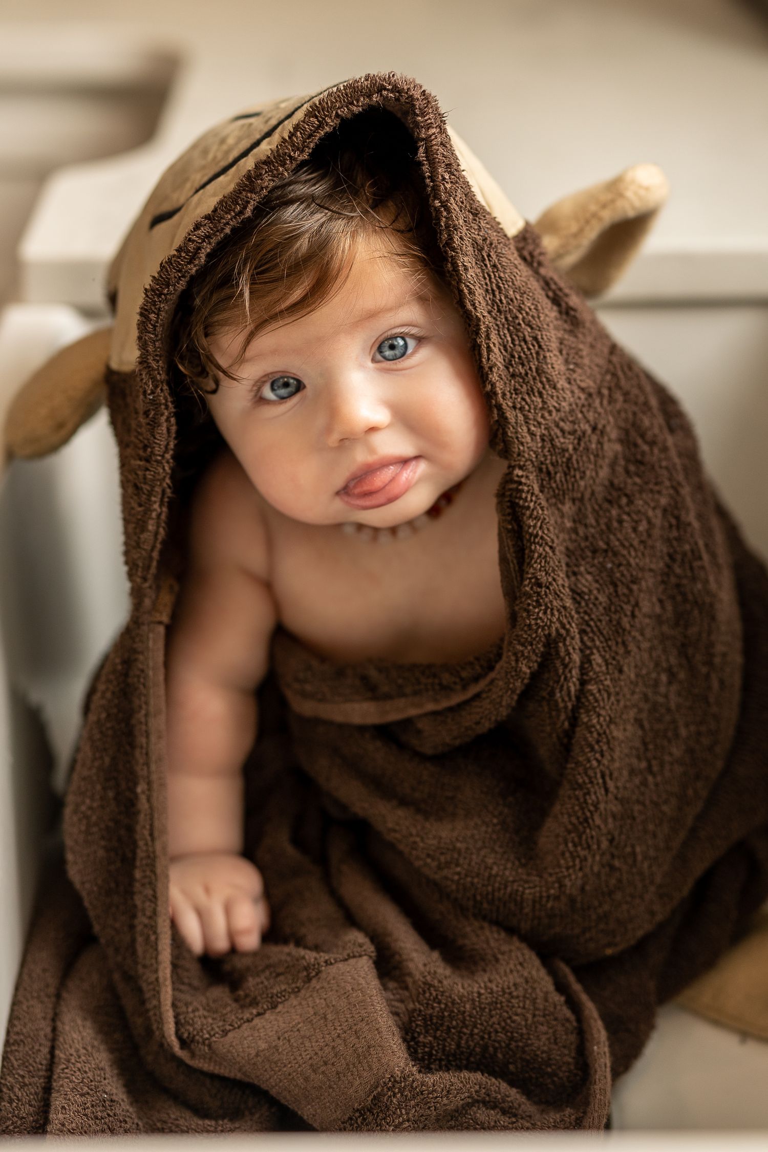 Adorable baby wrapped in a brown hooded animal towel with blue eyes and tongue out after bath time.