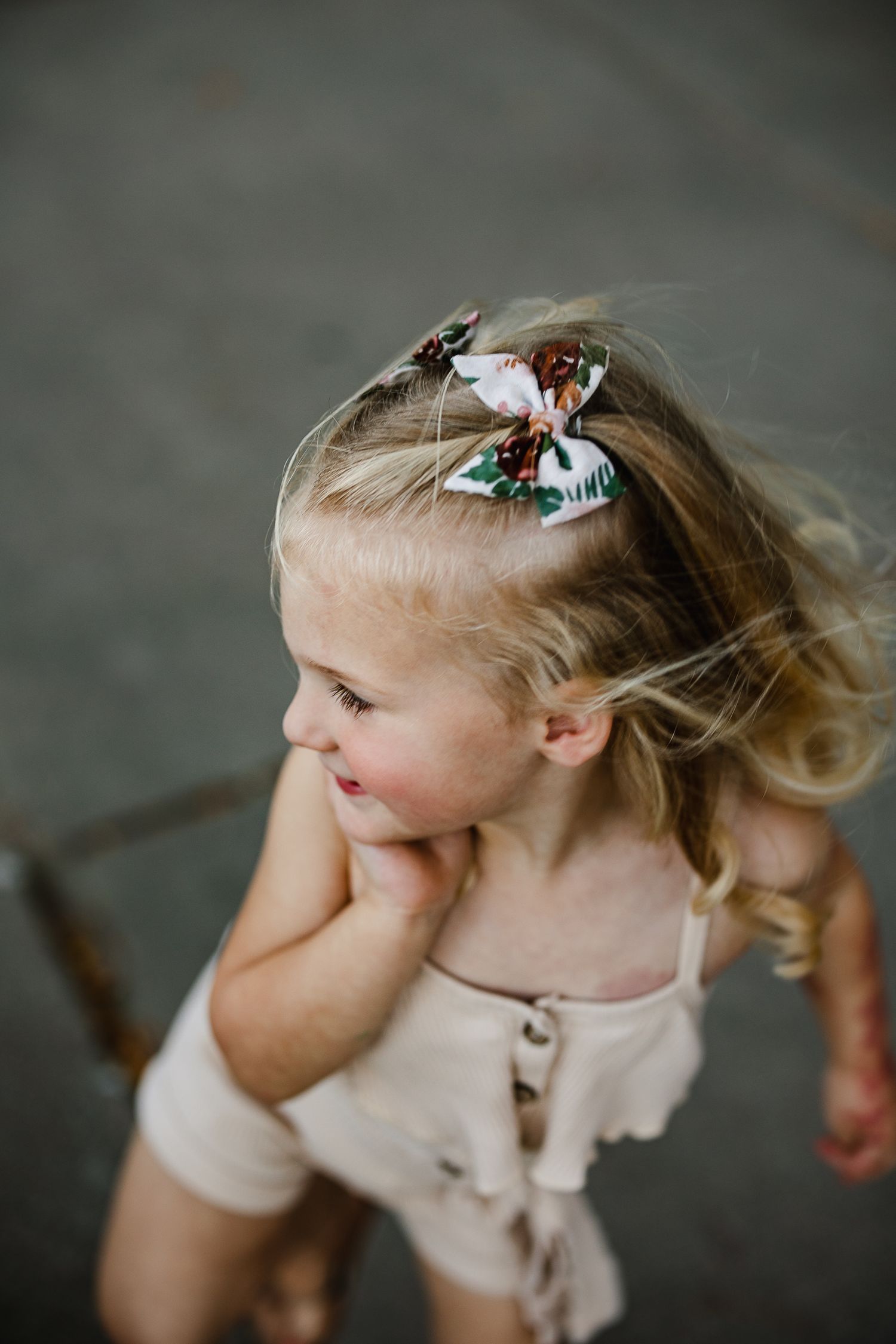 A young blonde toddler girl wearing a floral hair bow and cream romper, looking away in a candid outdoor photo.