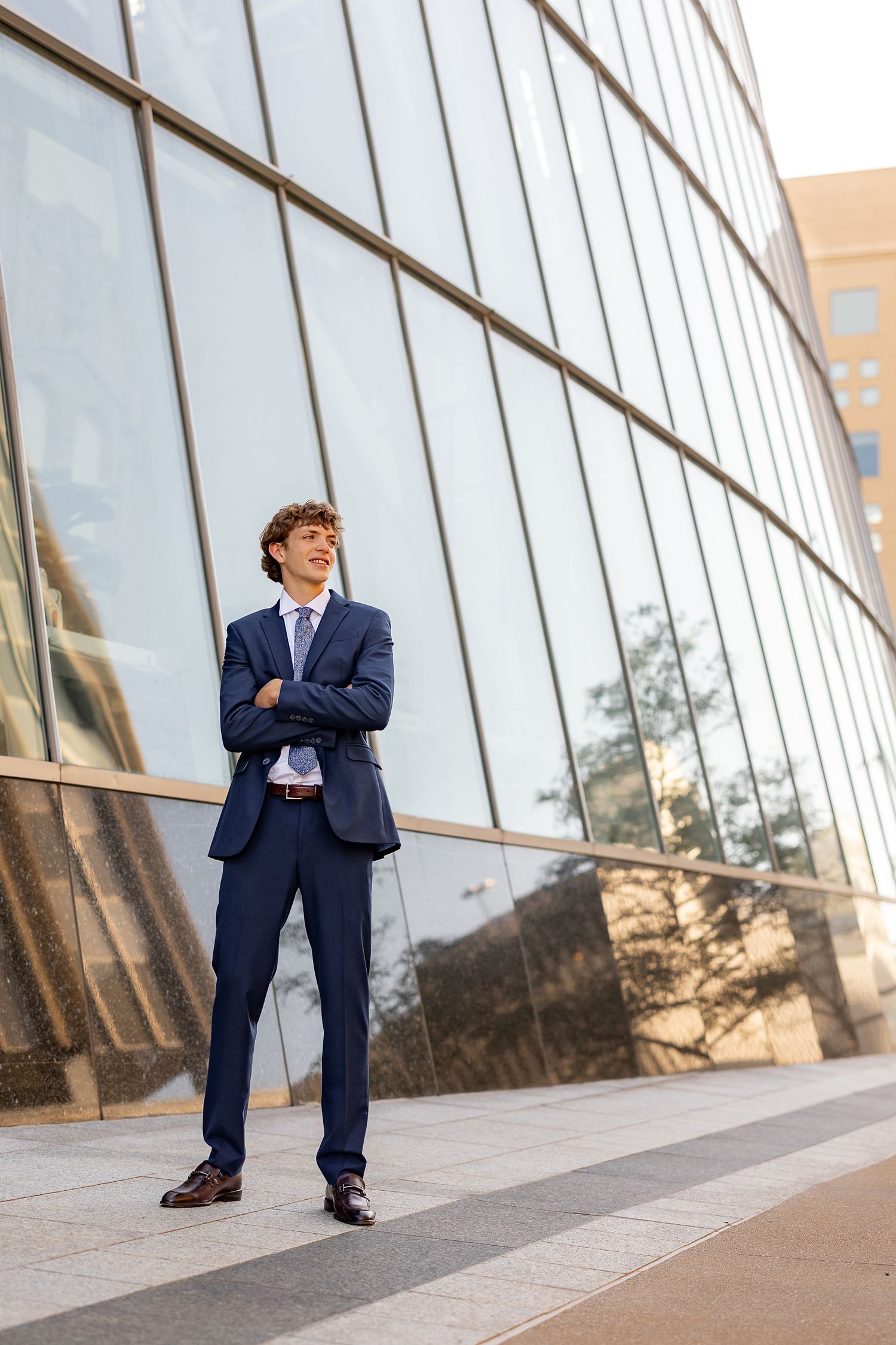 Young businessman in navy suit stands confidently with arms crossed outside modern glass office building.