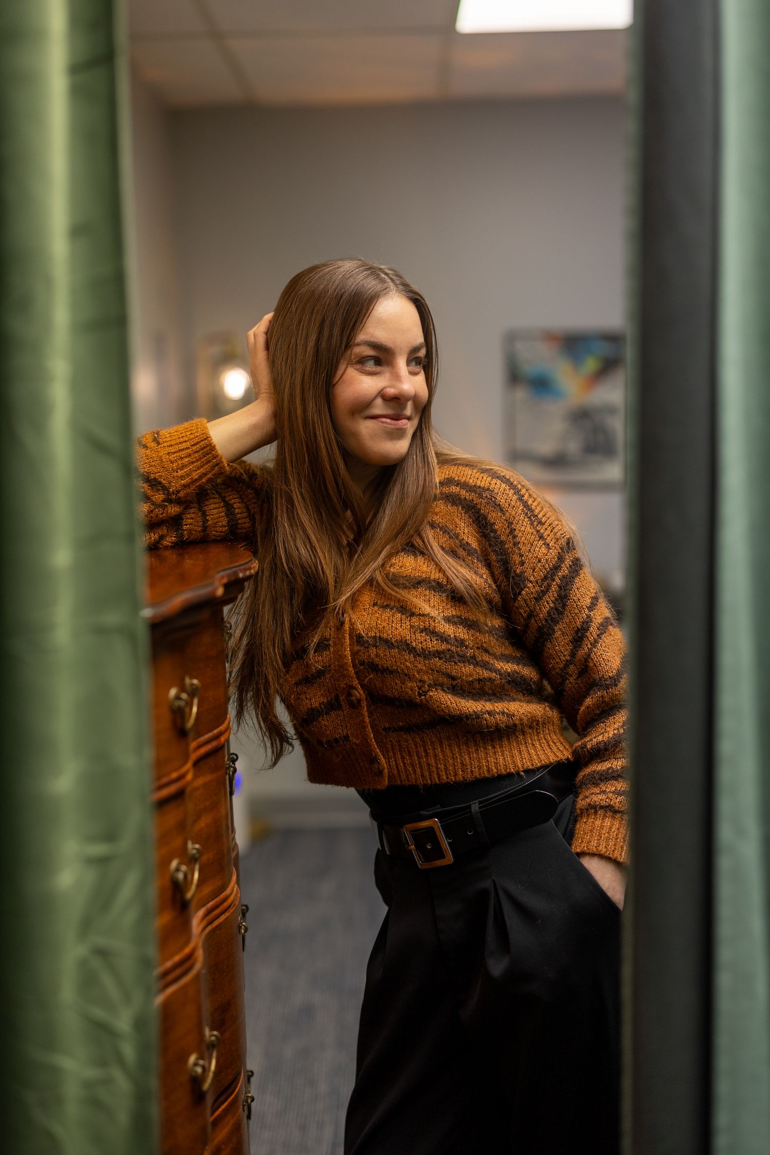 Smiling woman in tiger-print sweater and black pants leans against wooden dresser in a fitting room.