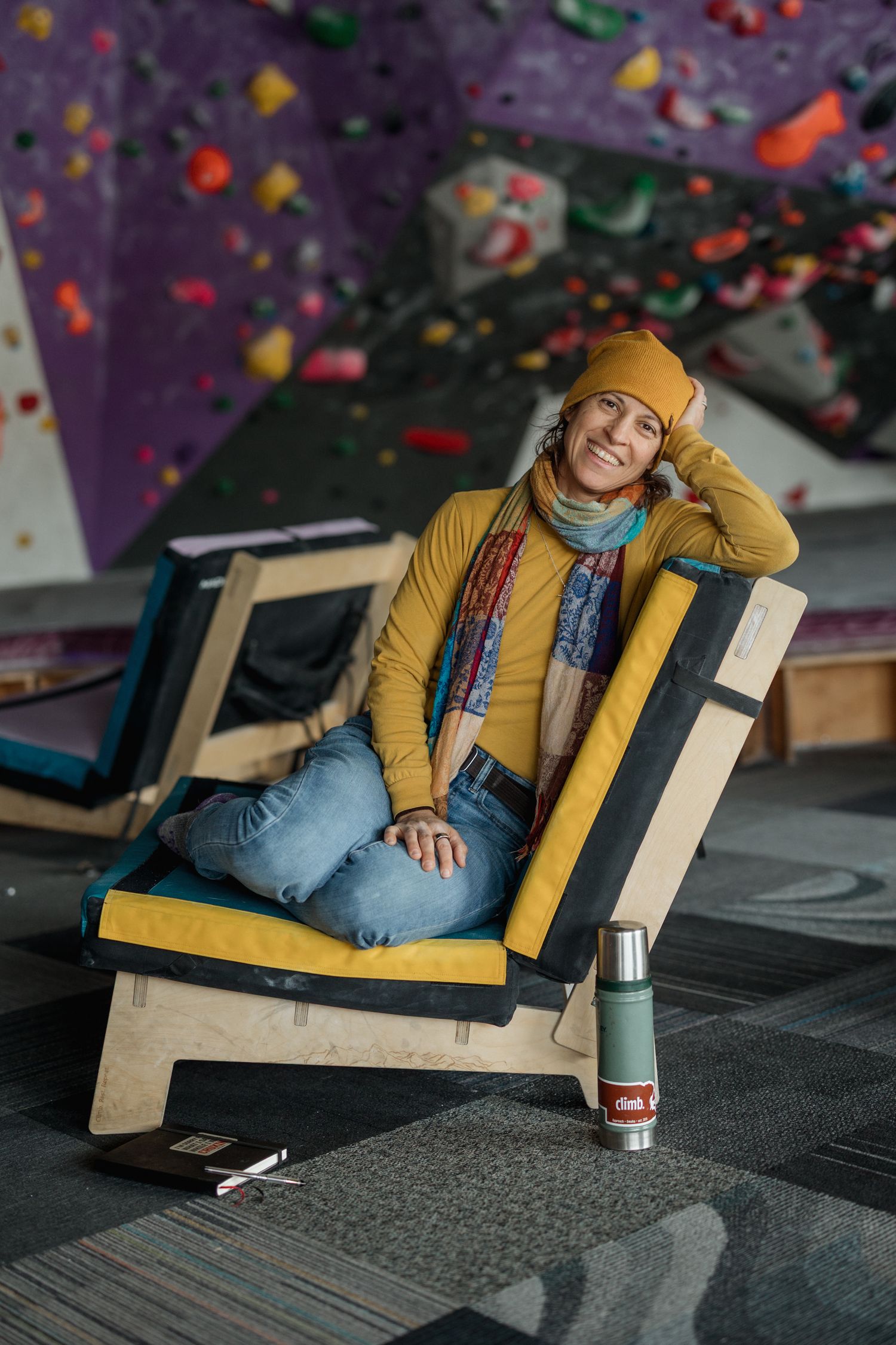 Smiling woman in yellow beanie and scarf sitting cross-legged in wooden chair at indoor climbing gym.