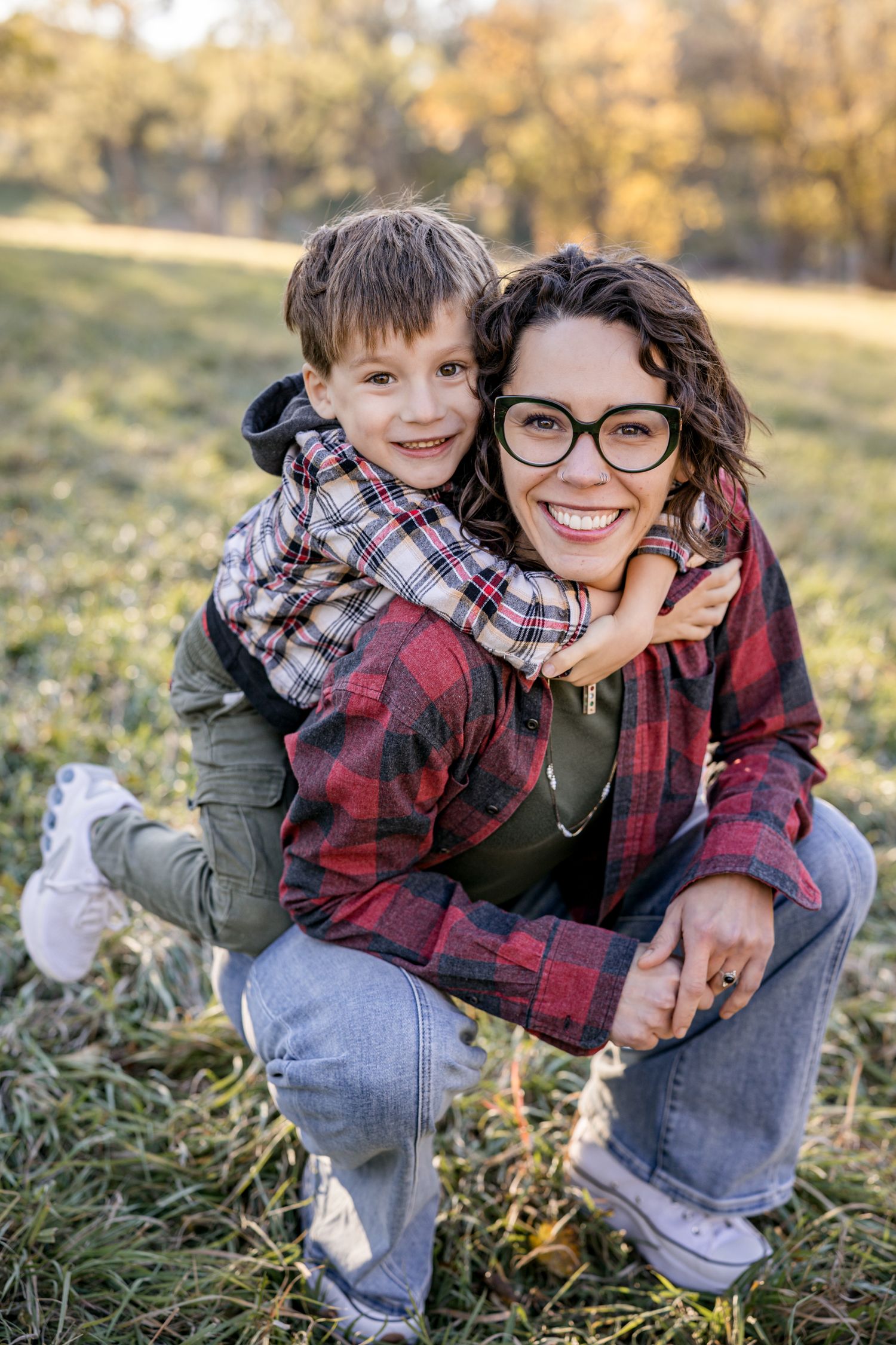 A smiling woman in plaid shirt crouches outdoors as a young boy hugs her from behind in a sunny autumn park.
