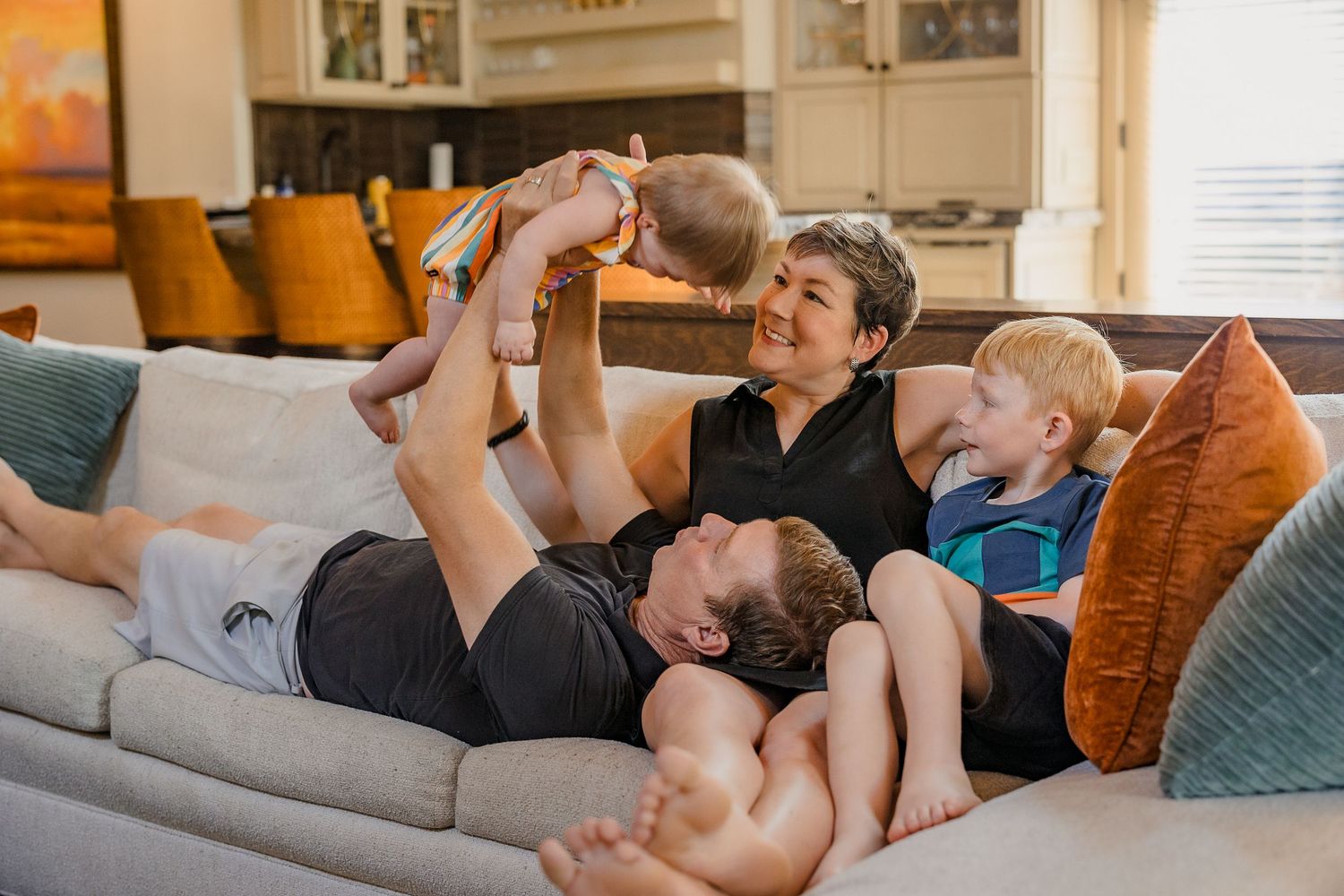 Happy family relaxing on a couch, parents playing with baby while young boy laughs in a cozy living room.