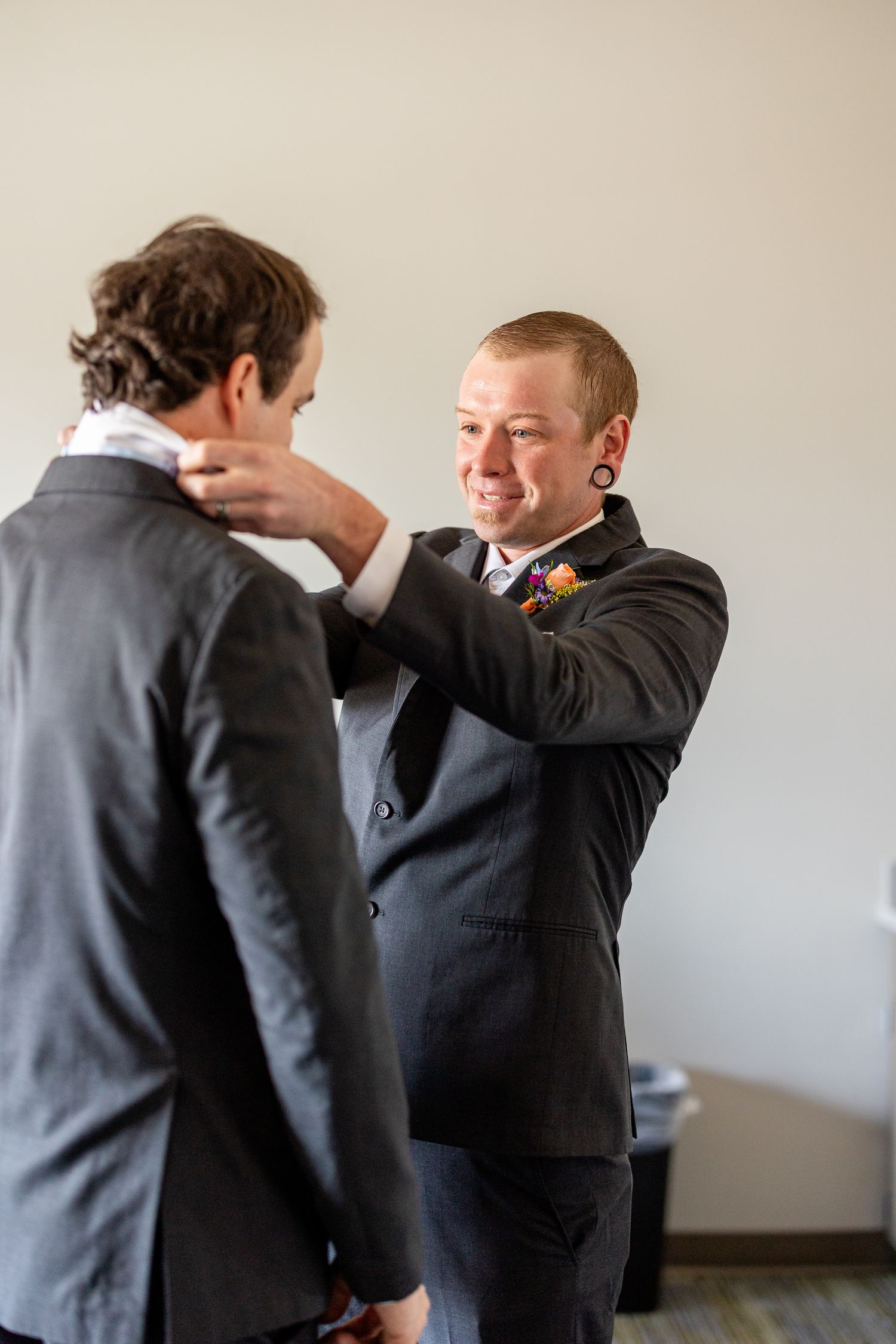 Groomsman smiling while adjusting the collar of another man in a dark suit at a wedding.