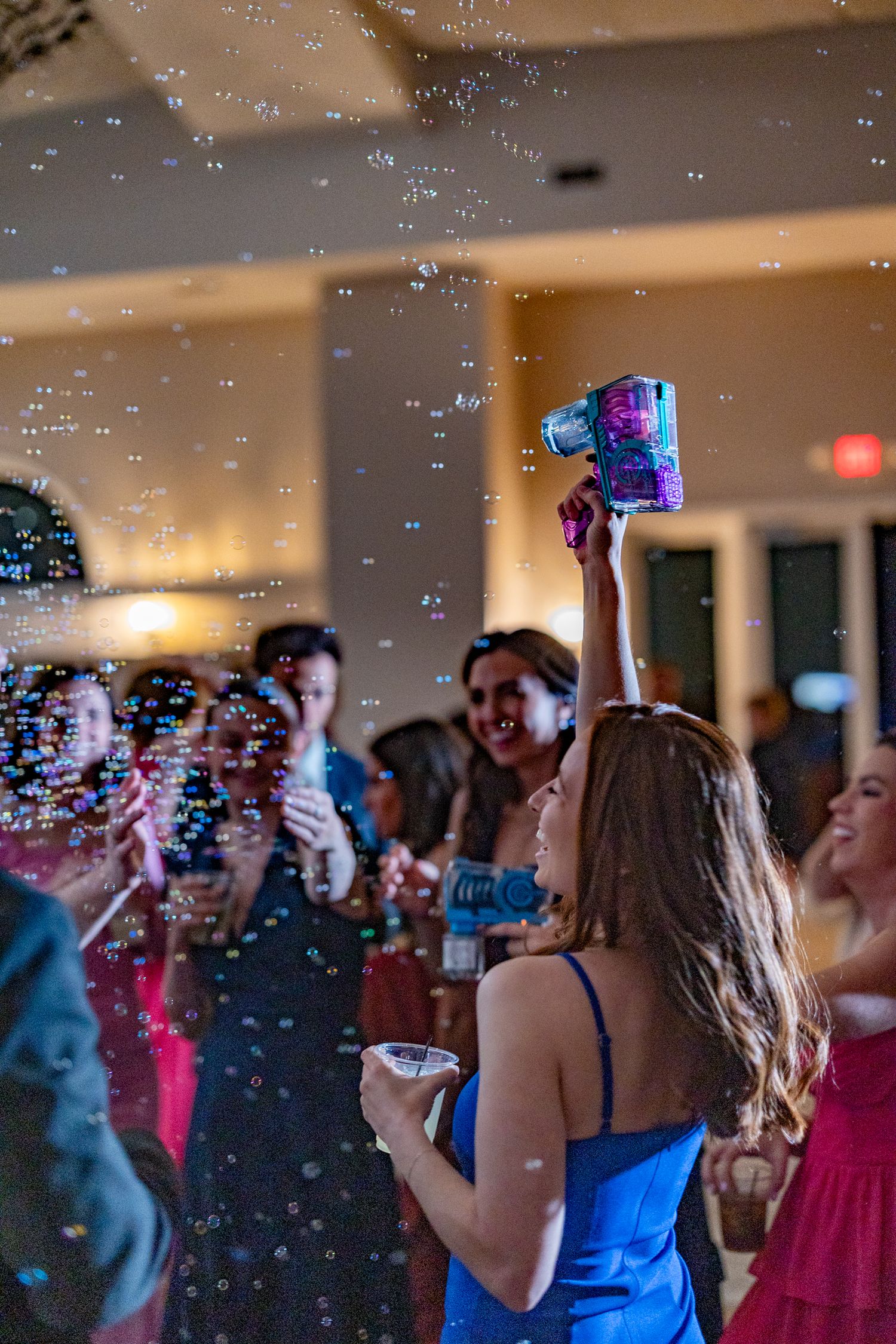 Woman in blue dress holds bubble machine aloft at a lively indoor party with guests dancing around her.